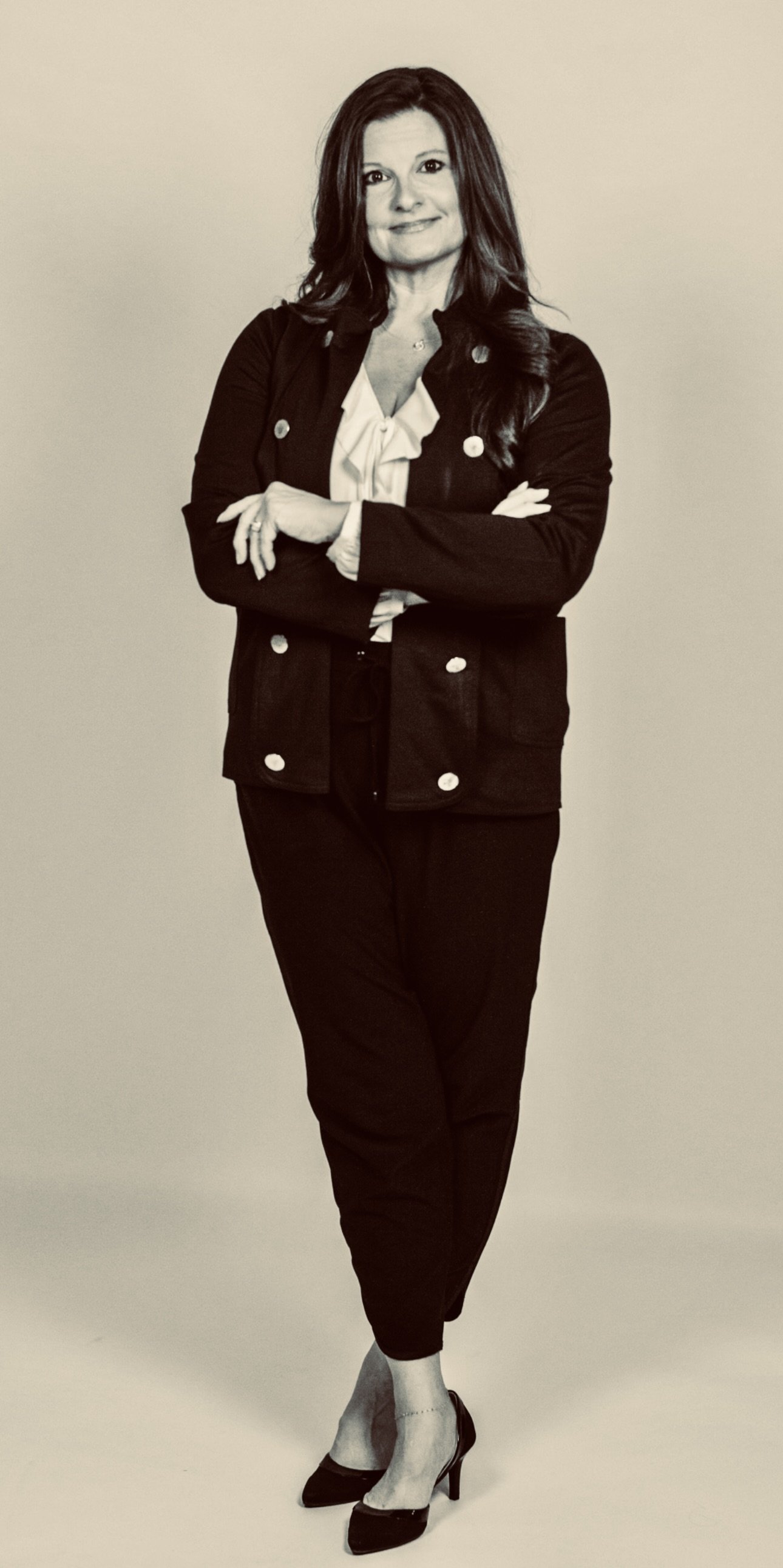 Woman with dark hair and a confident smile, wearing a blazer, white blouse, dark pants, and high heels, standing with arms crossed against a plain background.