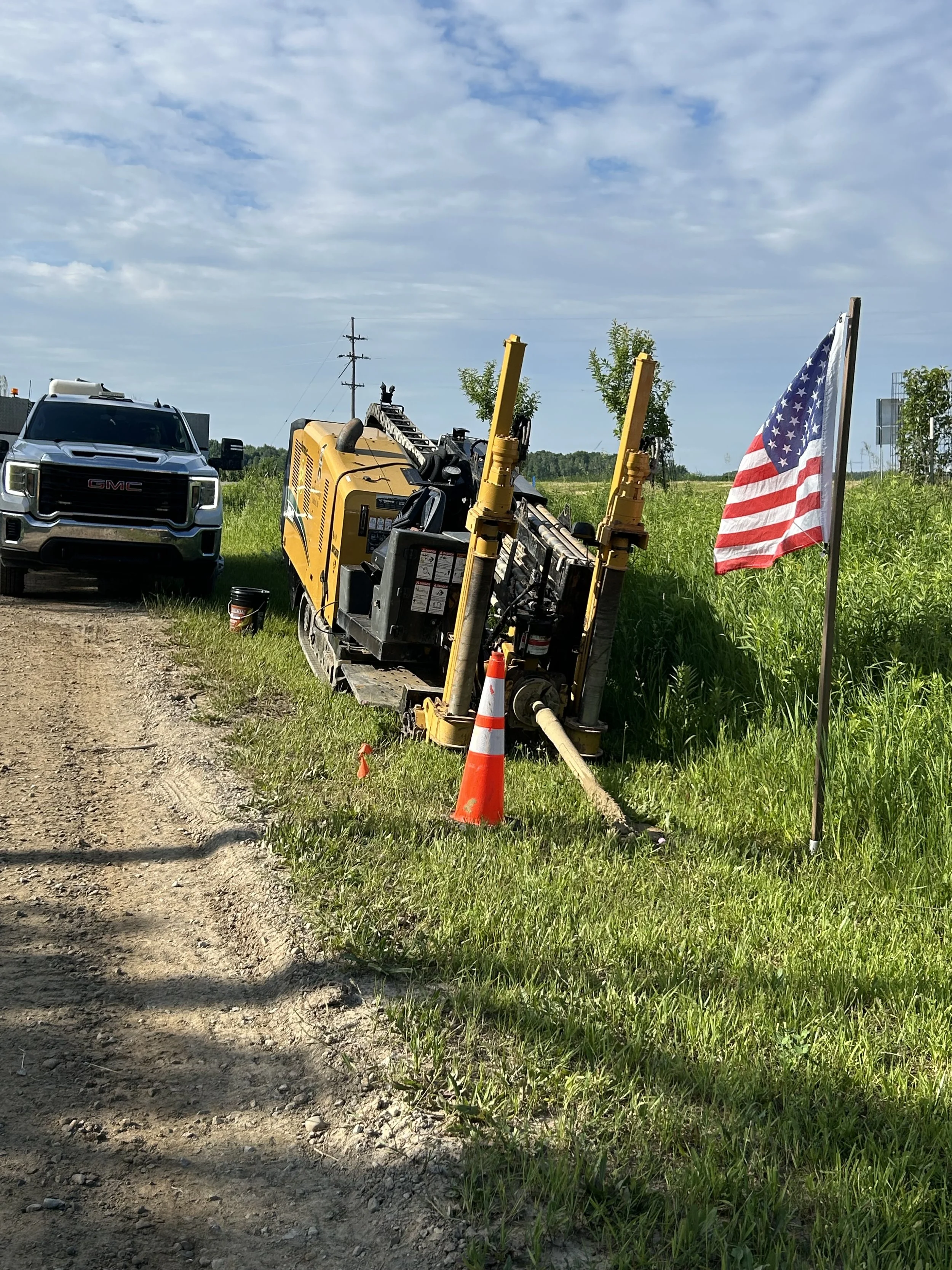 A capped construction site with a GMC pickup truck, a yellow drilling machine, orange safety cones, and an American flag on a grassy area under a partly cloudy sky.