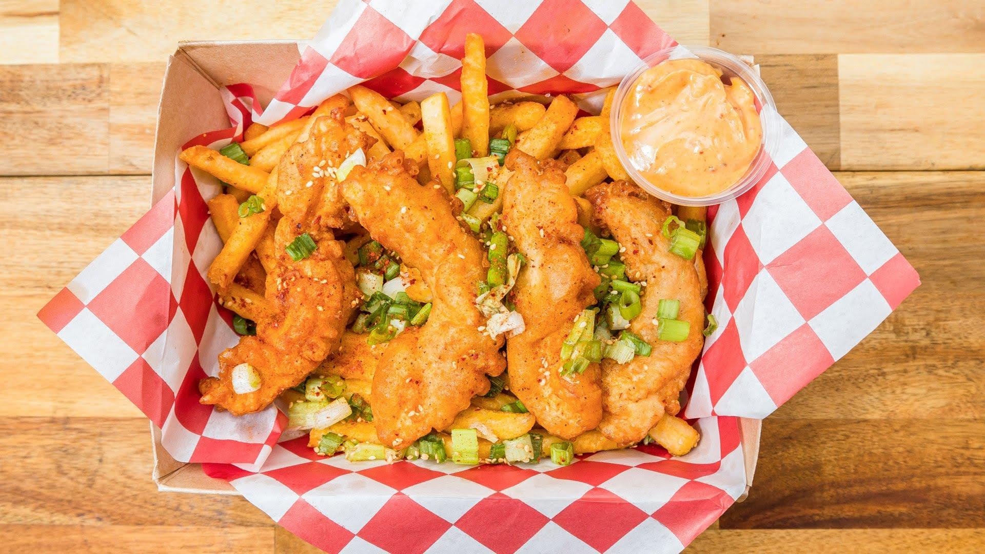 Basket of French fries topped with fried chicken tenders, chopped green onions, sesame seeds, and a side of creamy dipping sauce on a wooden table.