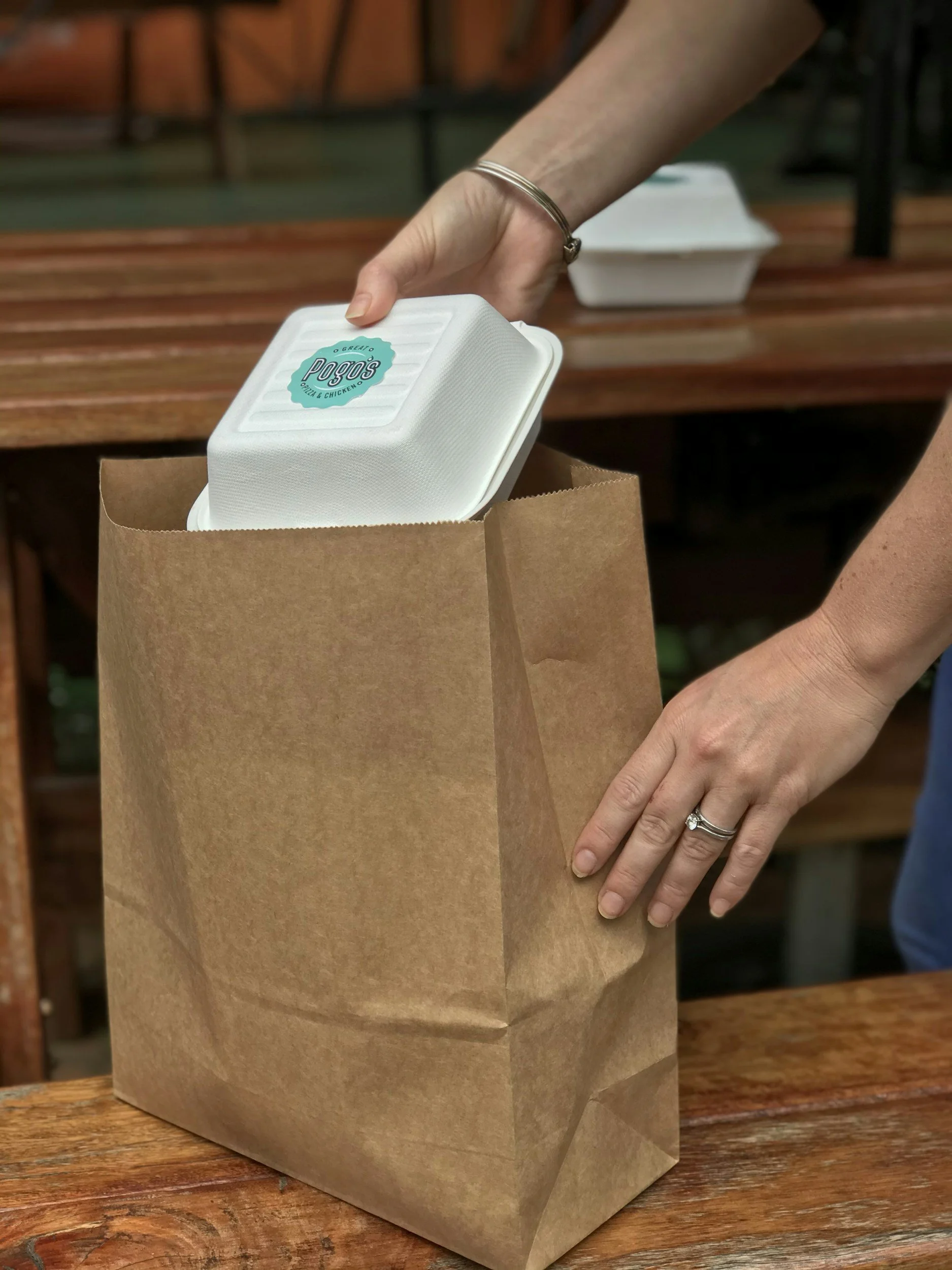 Person placing a takeout container into a brown paper bag on a wooden table.
