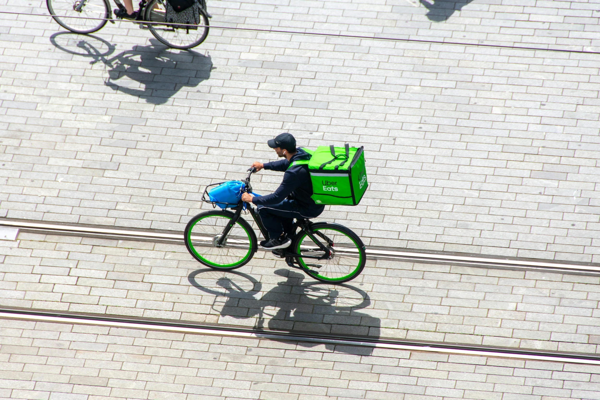 A person riding a bicycle with a green Uber Eats delivery bag on their back and a blue bag in the front basket, seen from above on a paved street with tram tracks.