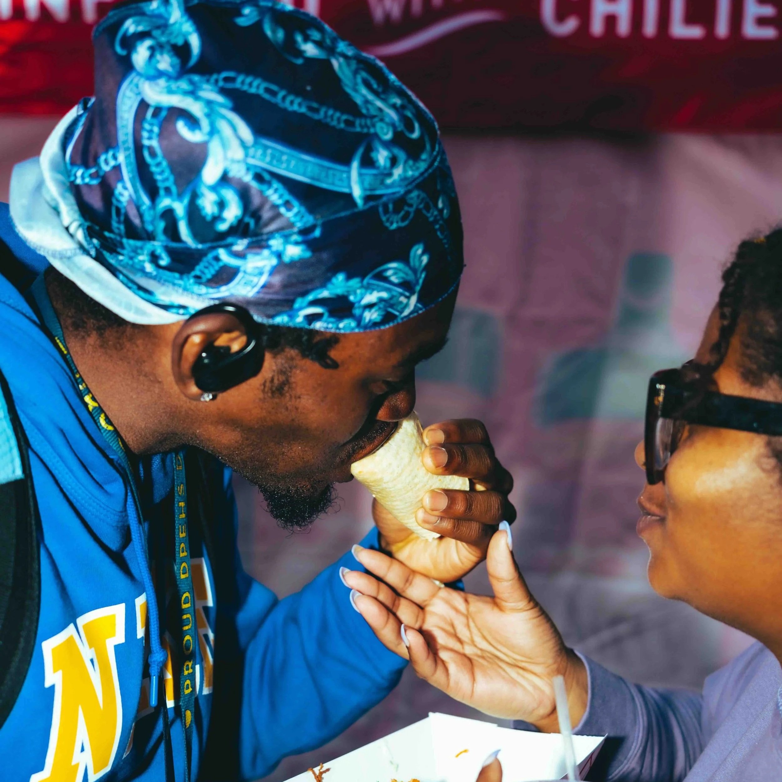 A man in a blue bandana and jacket is leaning forward with his mouth on an ice cream cone held by a woman with glasses, who is helping him eat it. They are close together, and the background includes a pink banner.