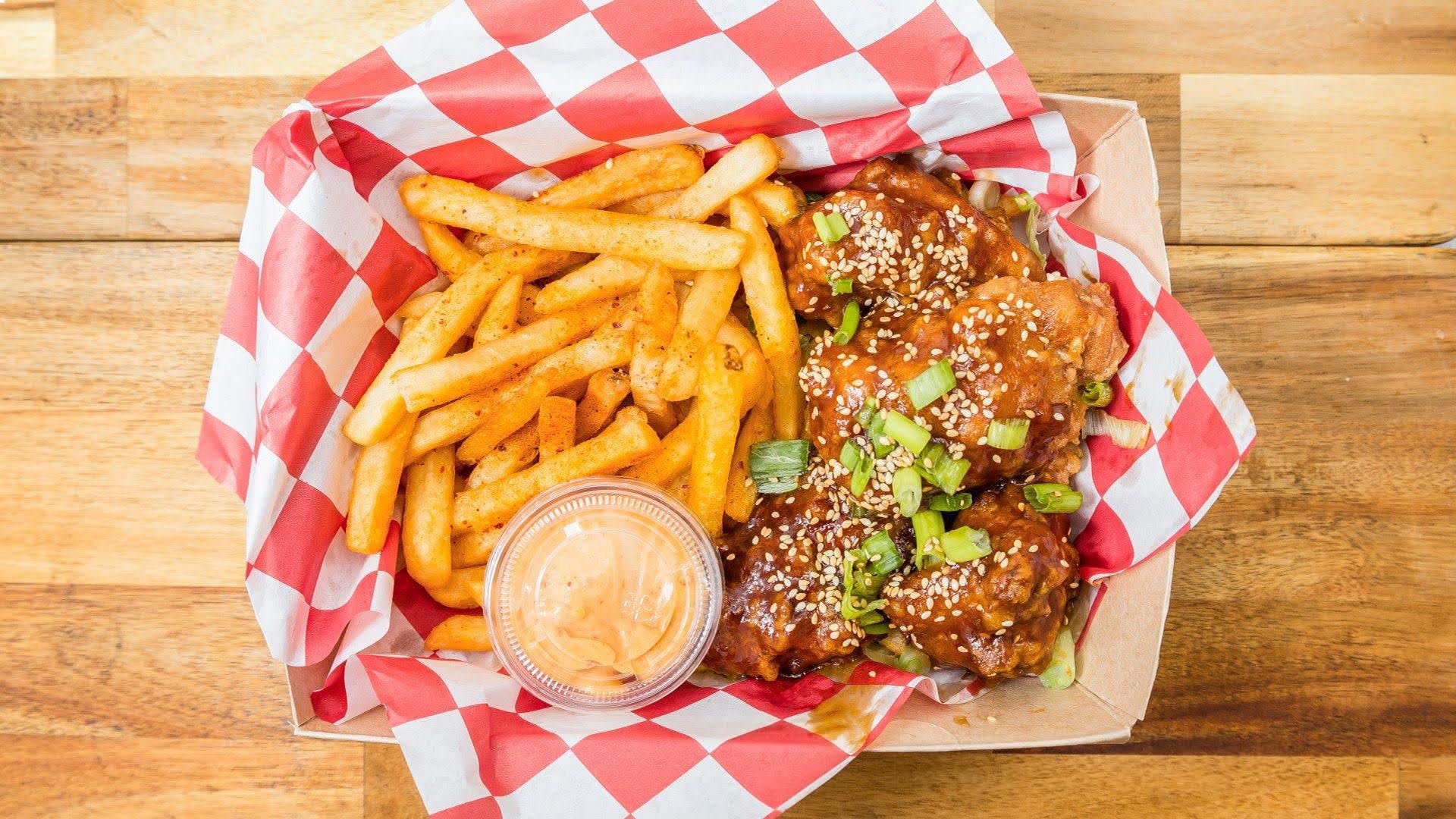 Basket with French fries, sauced chicken wings garnished with green onions and sesame seeds, and a small container of dipping sauce, on a wooden table.