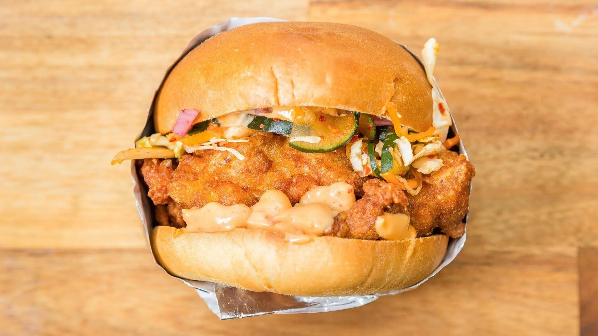 Close-up of a fried chicken sandwich with pickles, coleslaw, and sauce in a bun, on a wooden surface.