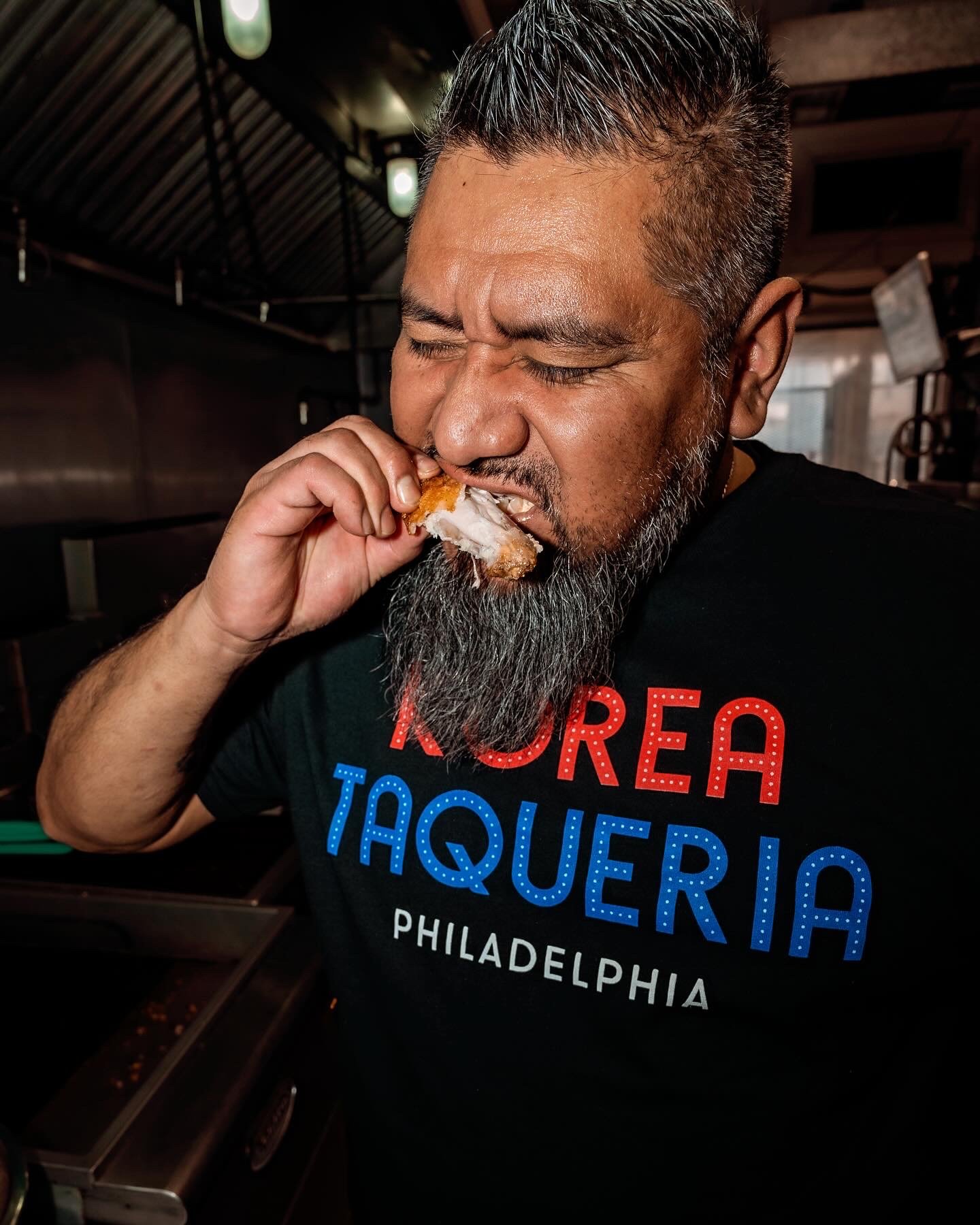 A man with a gray beard and gray hair is biting into a piece of fried chicken.