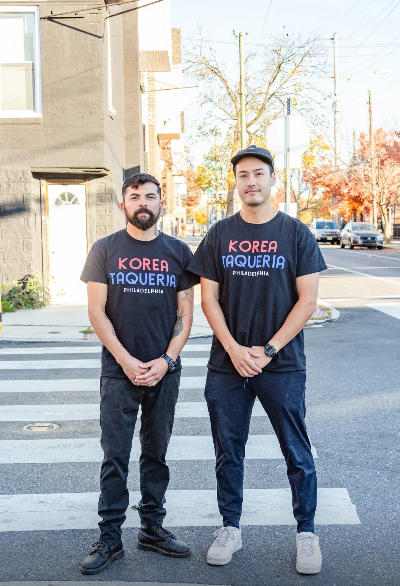 Two men standing on a crosswalk sidewalk in a city street during fall, wearing matching black T-shirts that say 'KOREA TAQUERIA PHILADELPHIA' with a background of colorful autumn trees and cars.