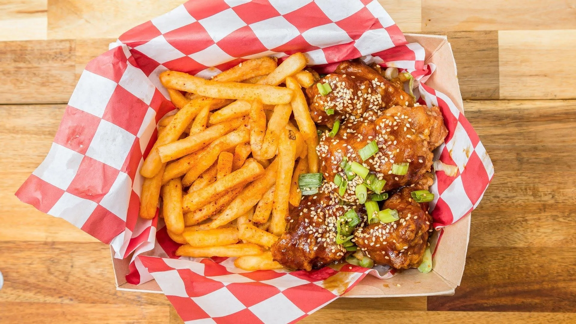 French fries and sauced chicken wings garnished with chopped green onions and sesame seeds, served in a takeout basket with red and white checkered paper on a wooden table.