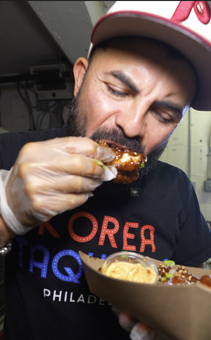 Man eating a glazed ribs with hand, holding a plate of food, wearing gloves and a baseball cap.