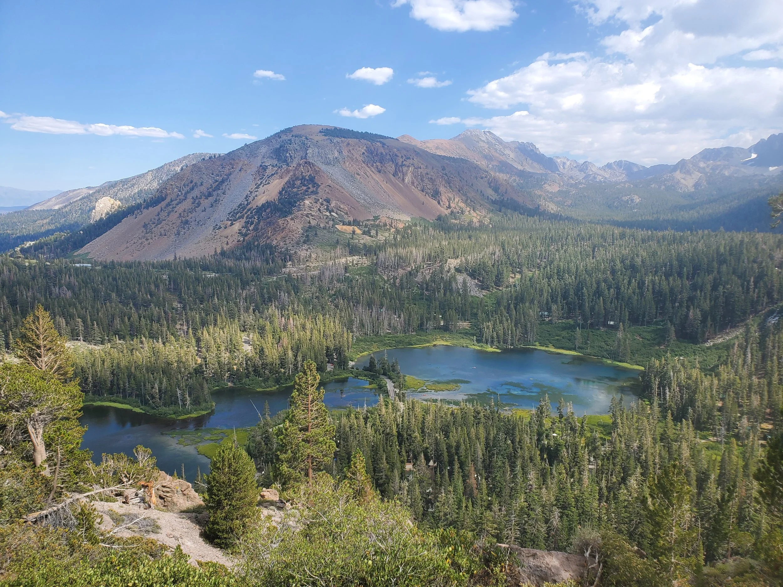 Mountain landscape with forested hills, a lake, and a partly cloudy sky.