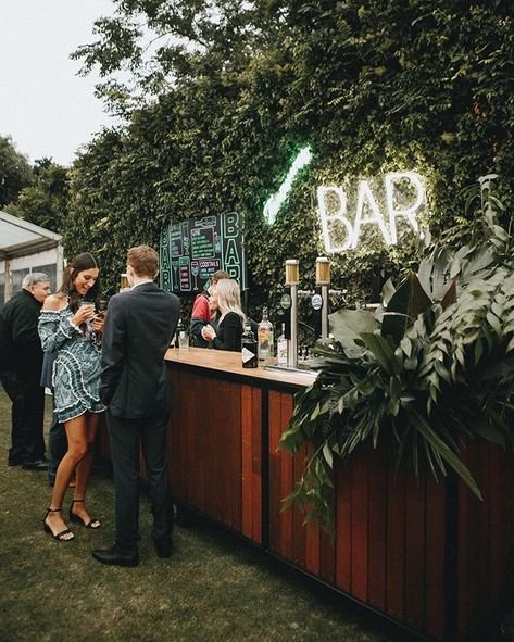 People gathered at an outdoor bar with a wooden counter, greenery, and a neon "BAR" sign.