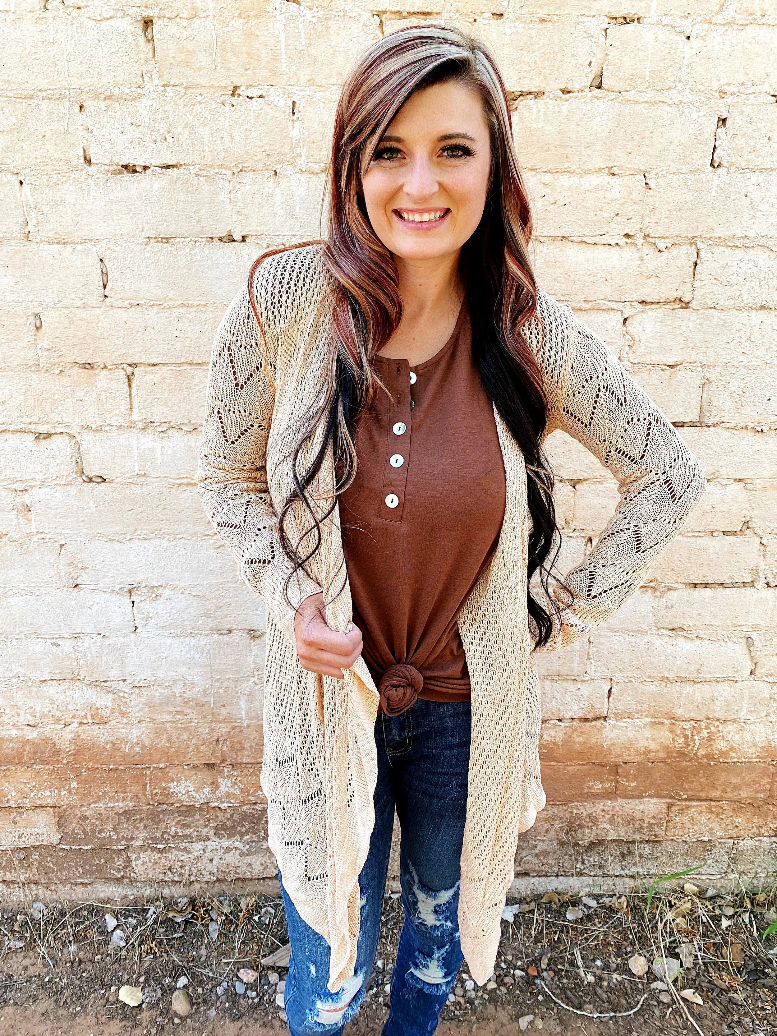 A young woman with long, dark wavy hair and a smile, standing outdoors against a light brick wall. She is wearing a brown top, a light-colored lace cardigan, and ripped jeans.