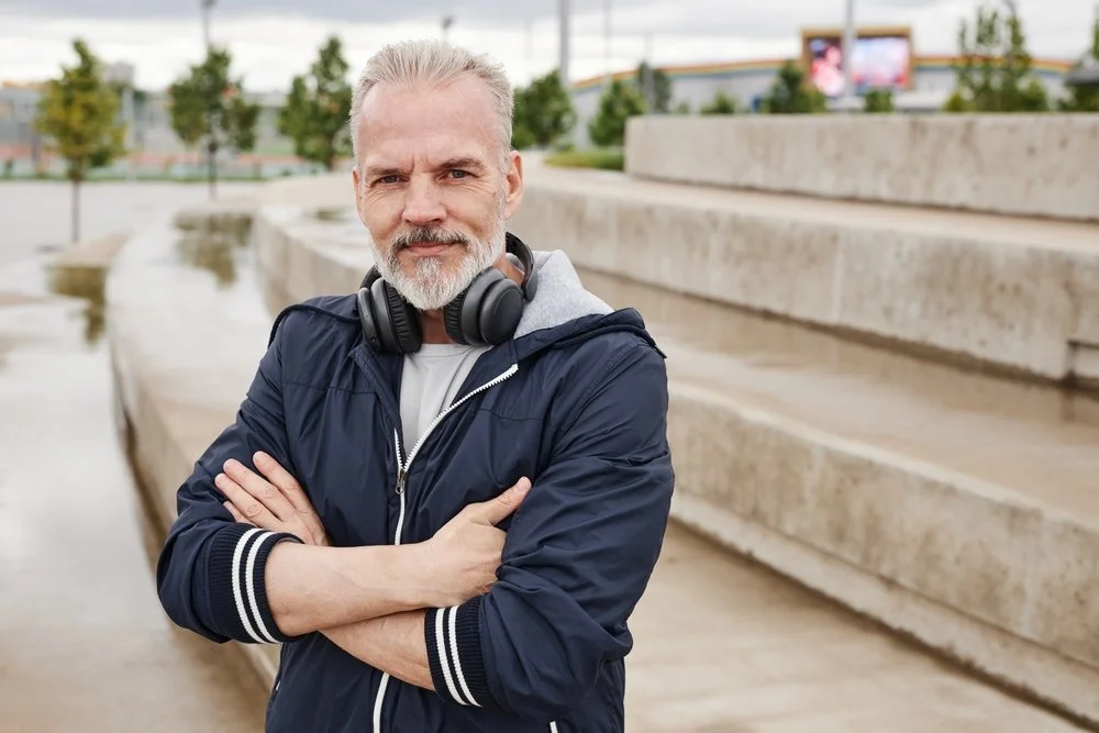 A confident man with gray hair and beard standing outdoors on steps, wearing a navy blue jacket and headphones around his neck.