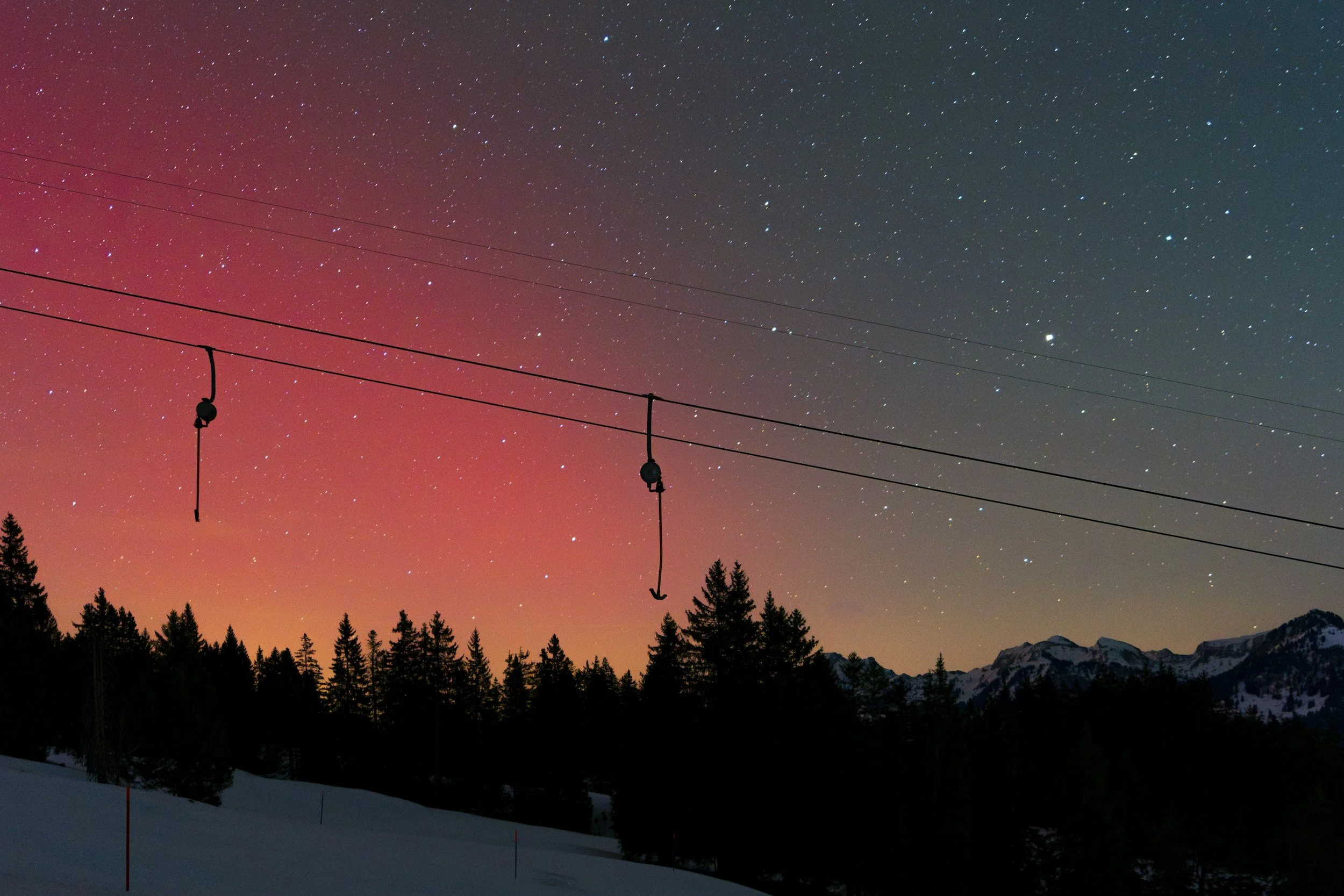 Night sky with stars and aurora, mountain silhouette, and ski lift cables with hooks.