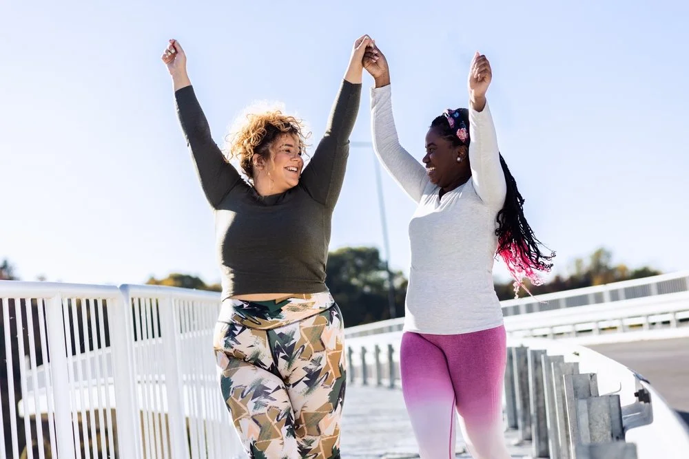 Two women happily holding hands and raising their arms in the air while walking outside on a bridge on a sunny day.