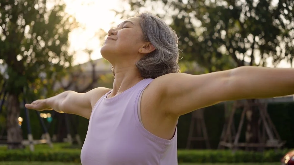 Older woman in a sleeveless top standing outdoors with arms outstretched, smiling and enjoying the sunlight.