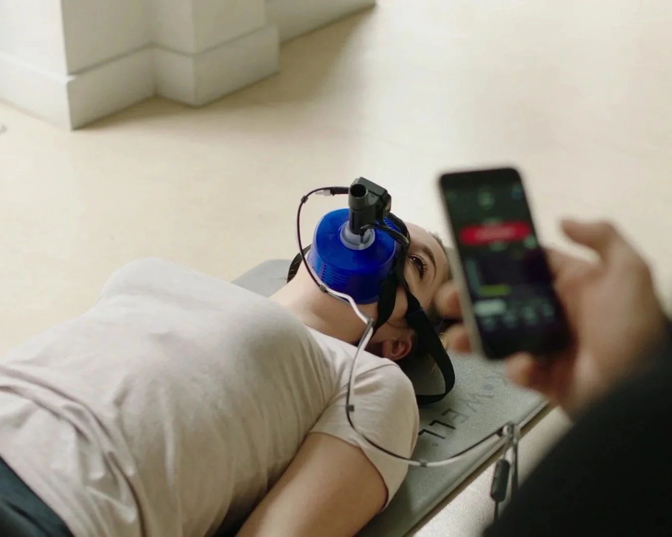 A person lying on a medical examination table with a head immobilizer, wearing a breathing mask connected to a ventilation device, while a medical professional prepares to record data with a handheld device.