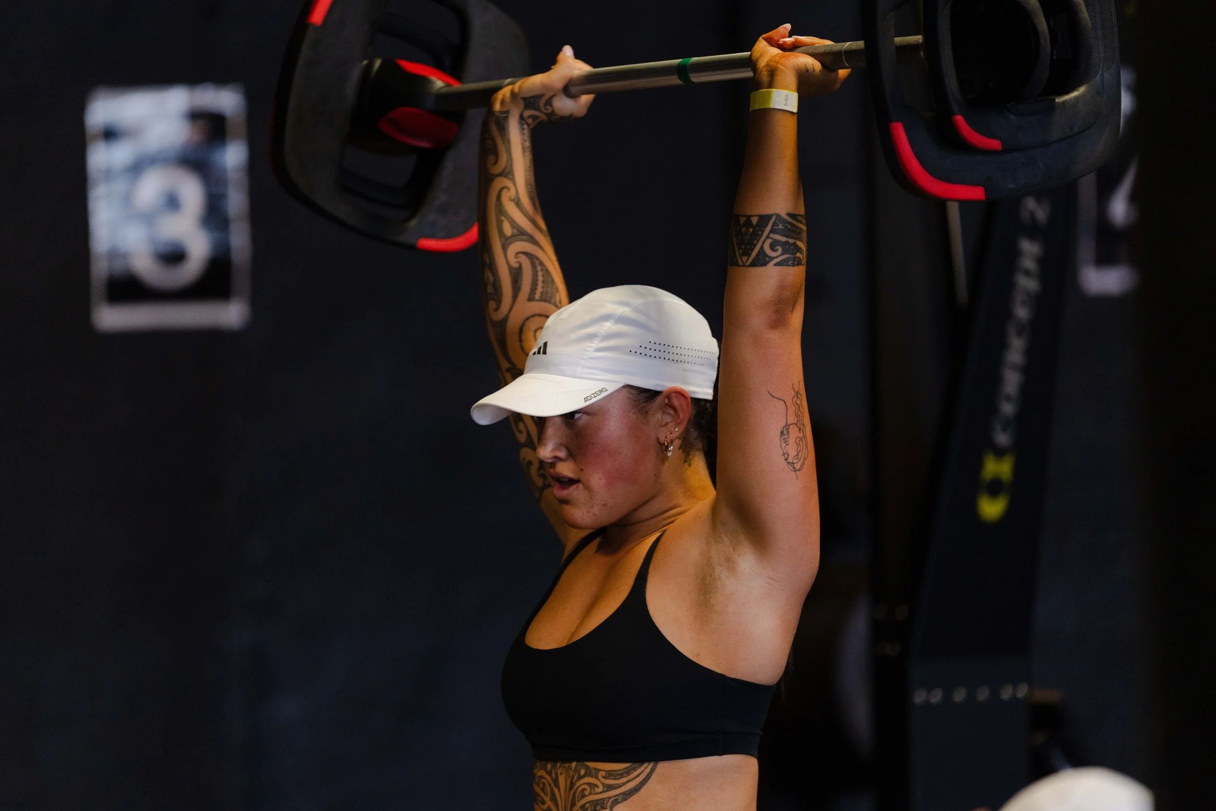A Maori woman wearing a white cap and black sports bra lifting a barbell overhead at Les Mills gym.