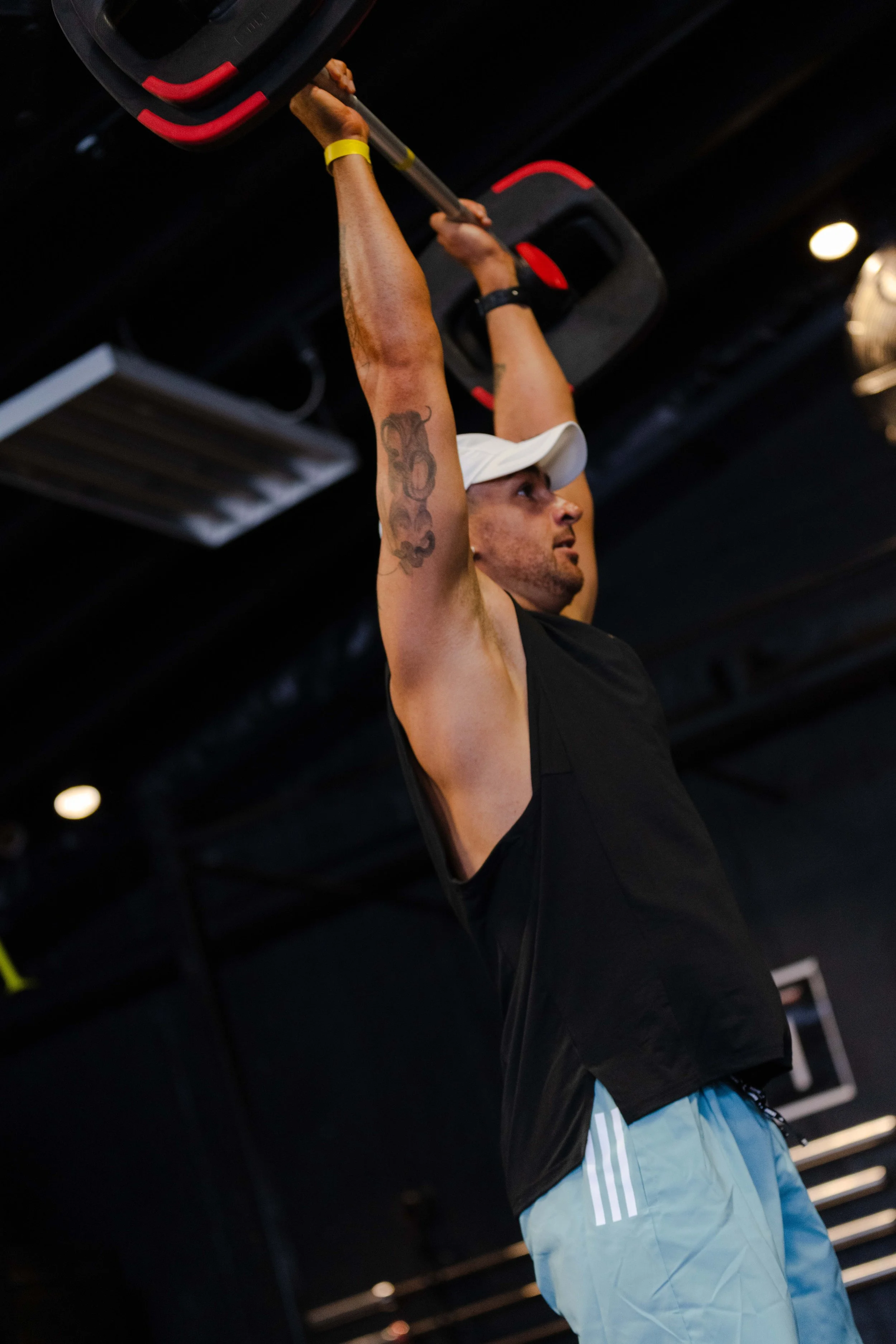 A man performing a pull-up exercise in a Les Mills gym, wearing a white cap, black sleeveless shirt, and light-colored shorts.