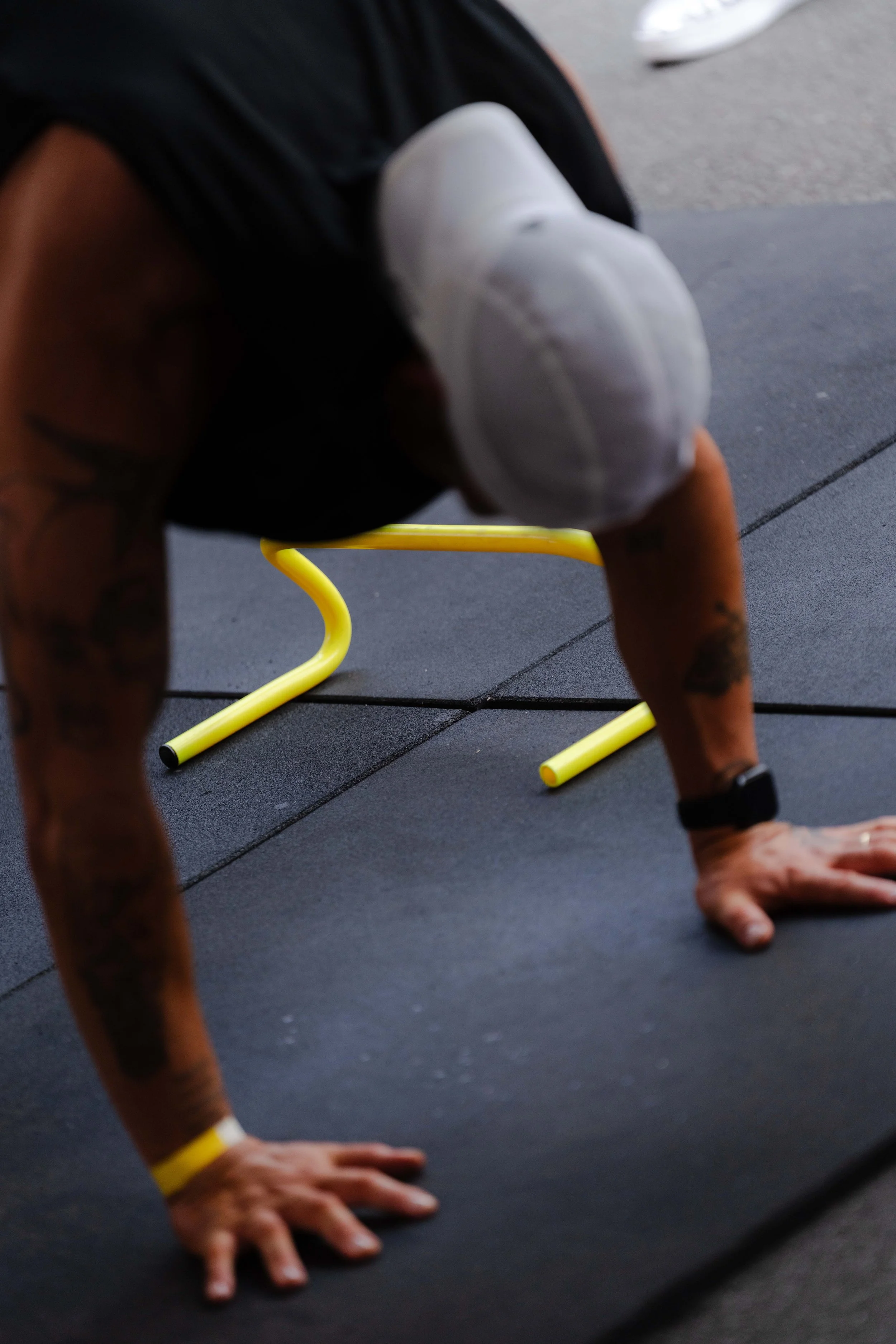 A person in a white hat and black shirt is doing push-ups on a black surface with yellow workout equipment in the background.