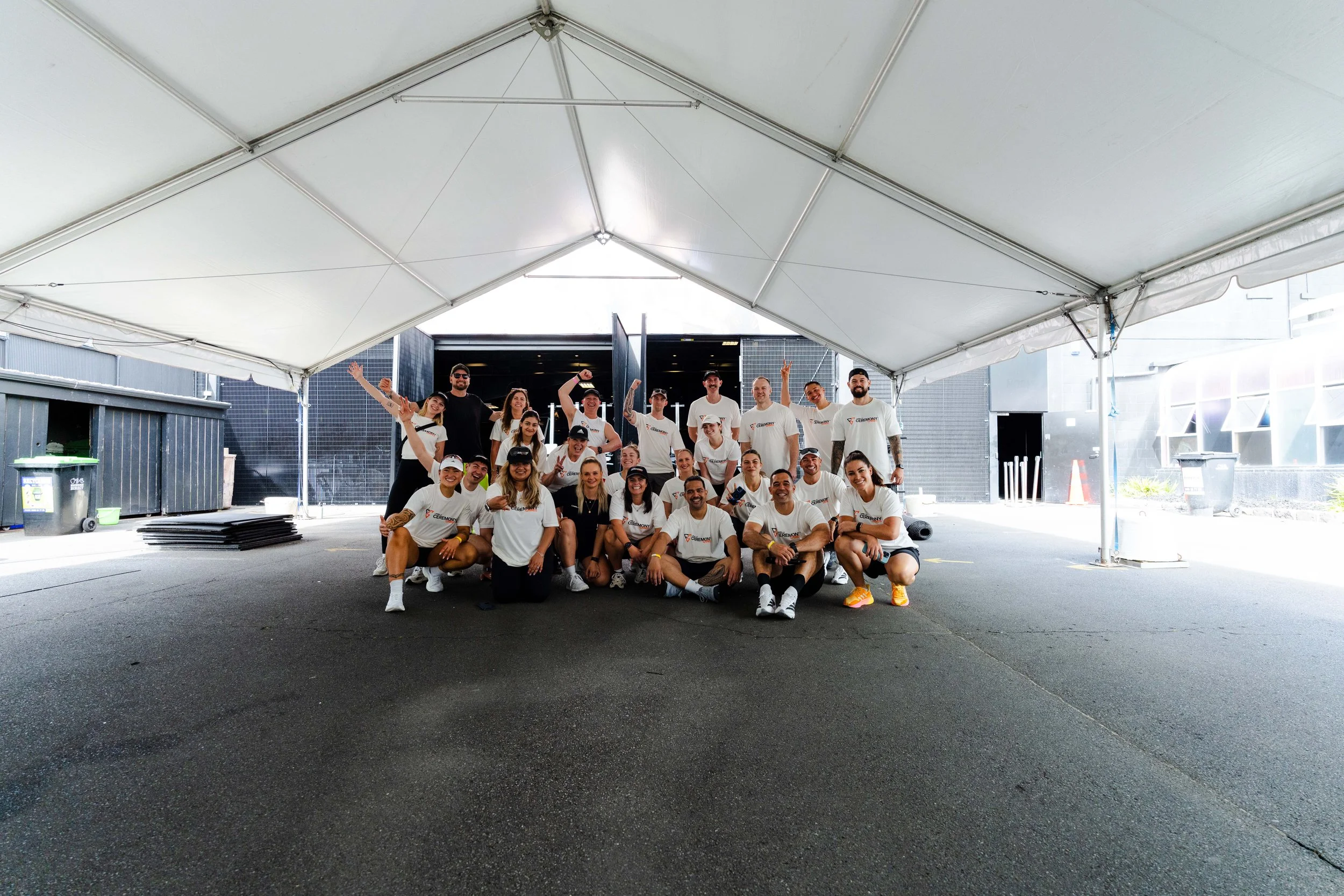 Group of people under a large white tent at Les Mills Ceremony Games in Wellington, some standing and some kneeling, smiling, wearing white T-shirts and caps.
