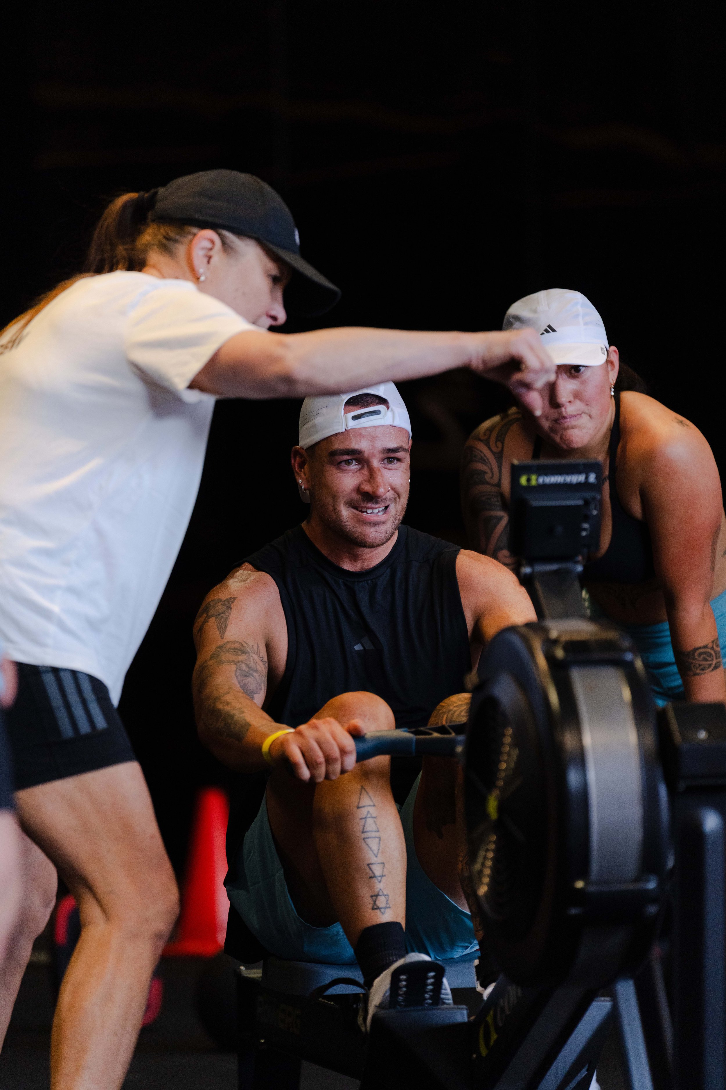 A man participating in an indoor rowing workout, with a woman coach and another woman assisting, all wearing athletic attire.