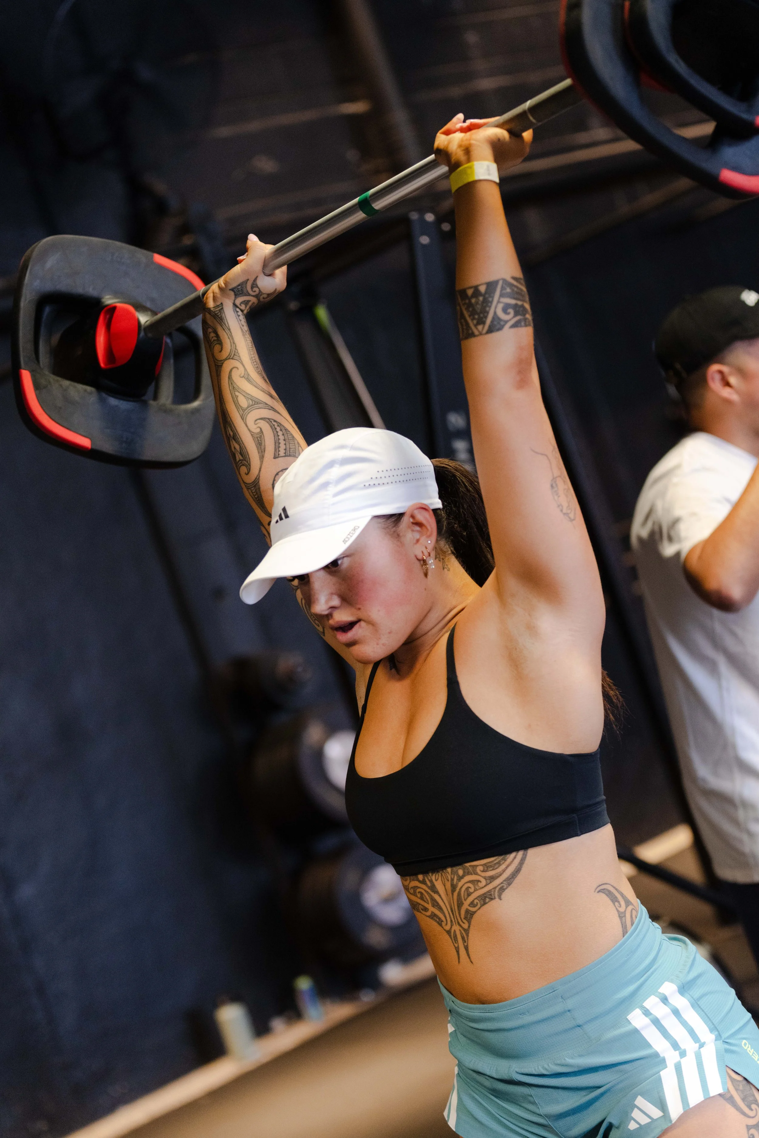 A Maori woman wearing a black sports bra, light blue shorts, a white cap, and tattoos on her arms and torso, is lifting a barbell overhead in a Les Mills gym.
