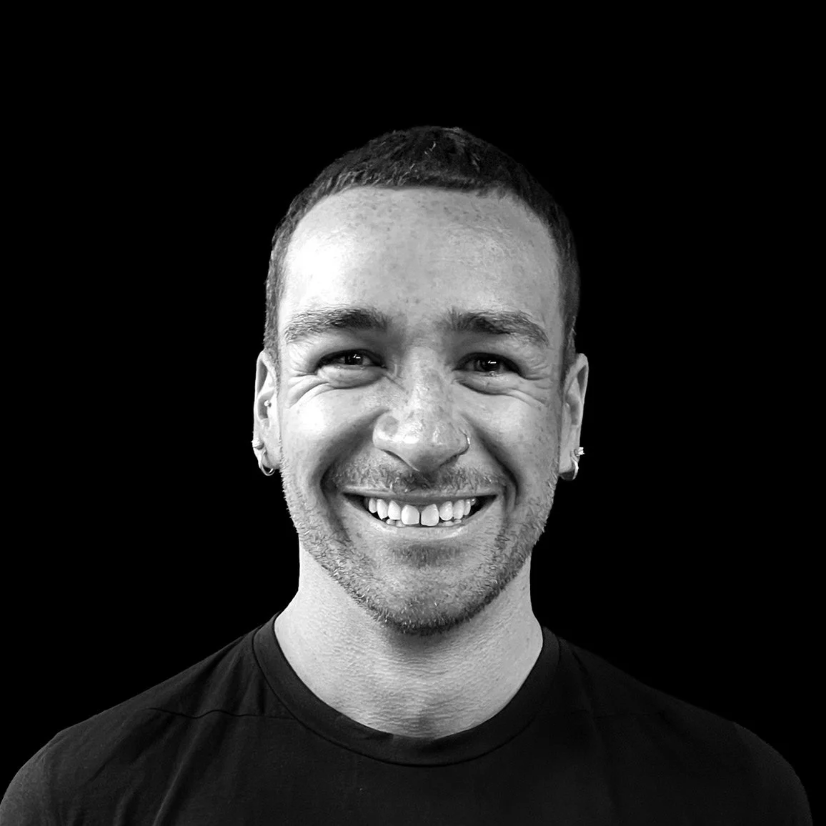 Black and white portrait of Nate Waghorn-Wright smiling man with short hair, earrings, and a nose piercing, wearing a black shirt, against a black background.