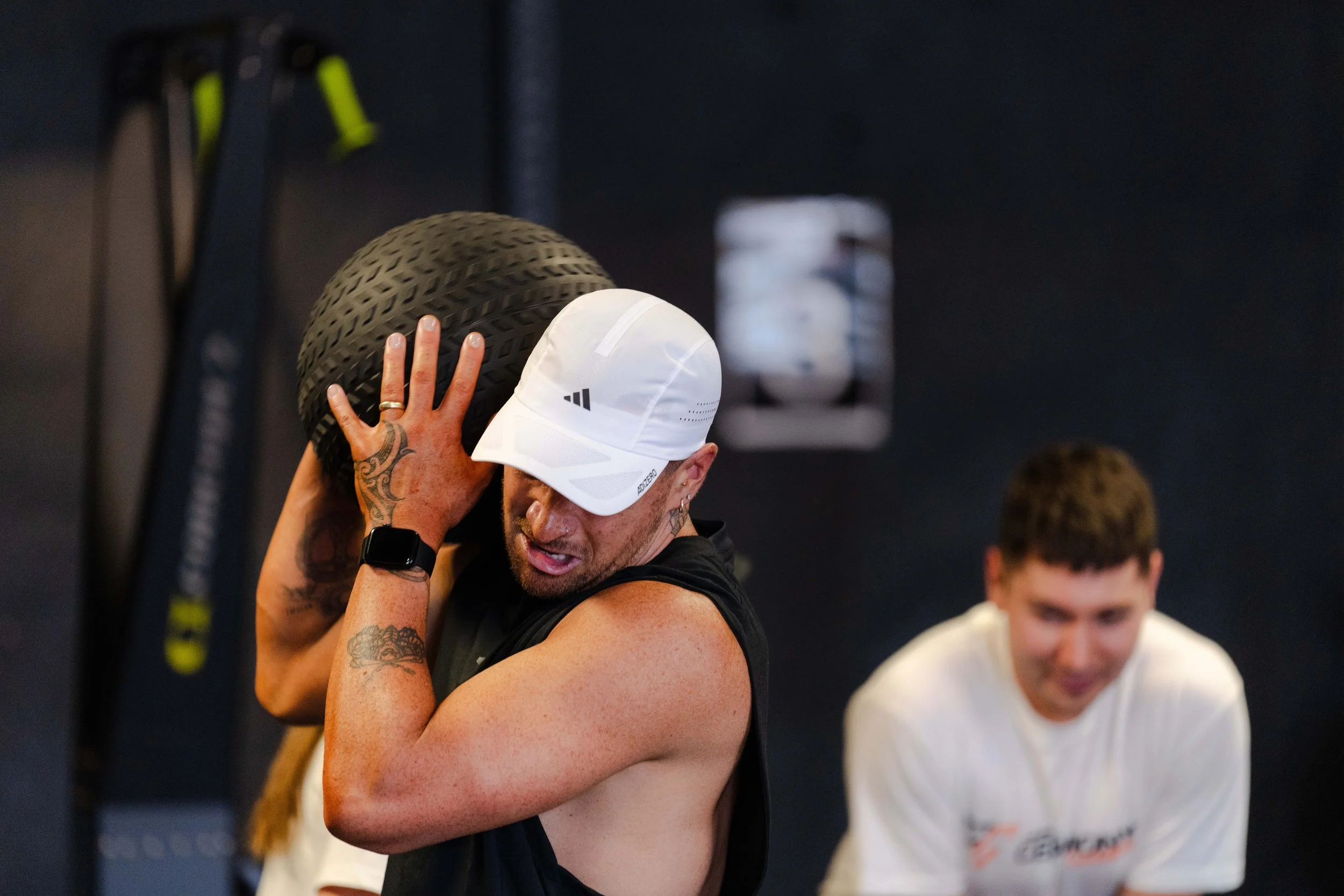 Man lifting a large black tire during a workout session in a gym, with another person blurred in the background.