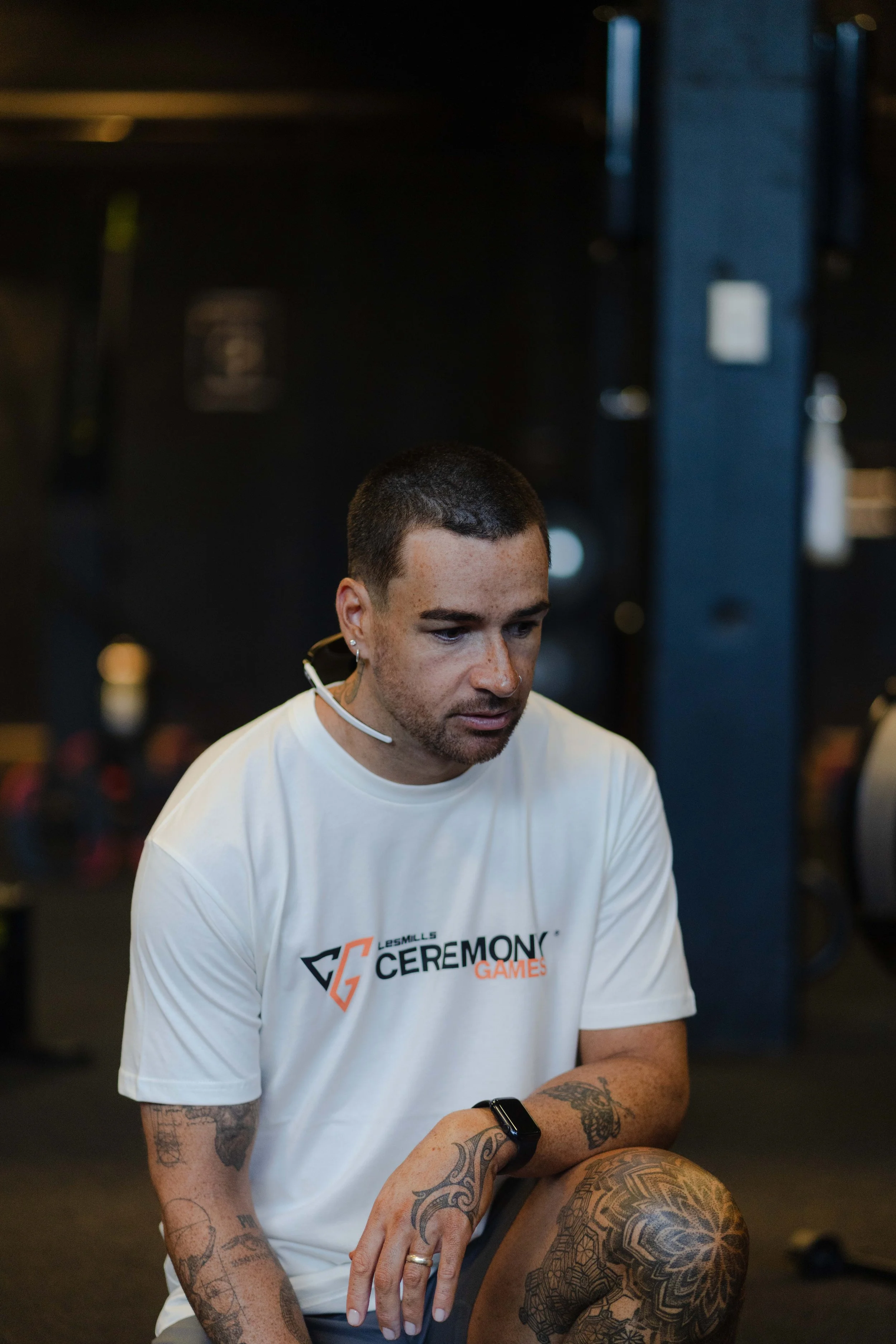 Young man with tattoos, wearing a white T-shirt with 'Les Mills Ceremonies Games' logo, sitting and looking down in a gym.