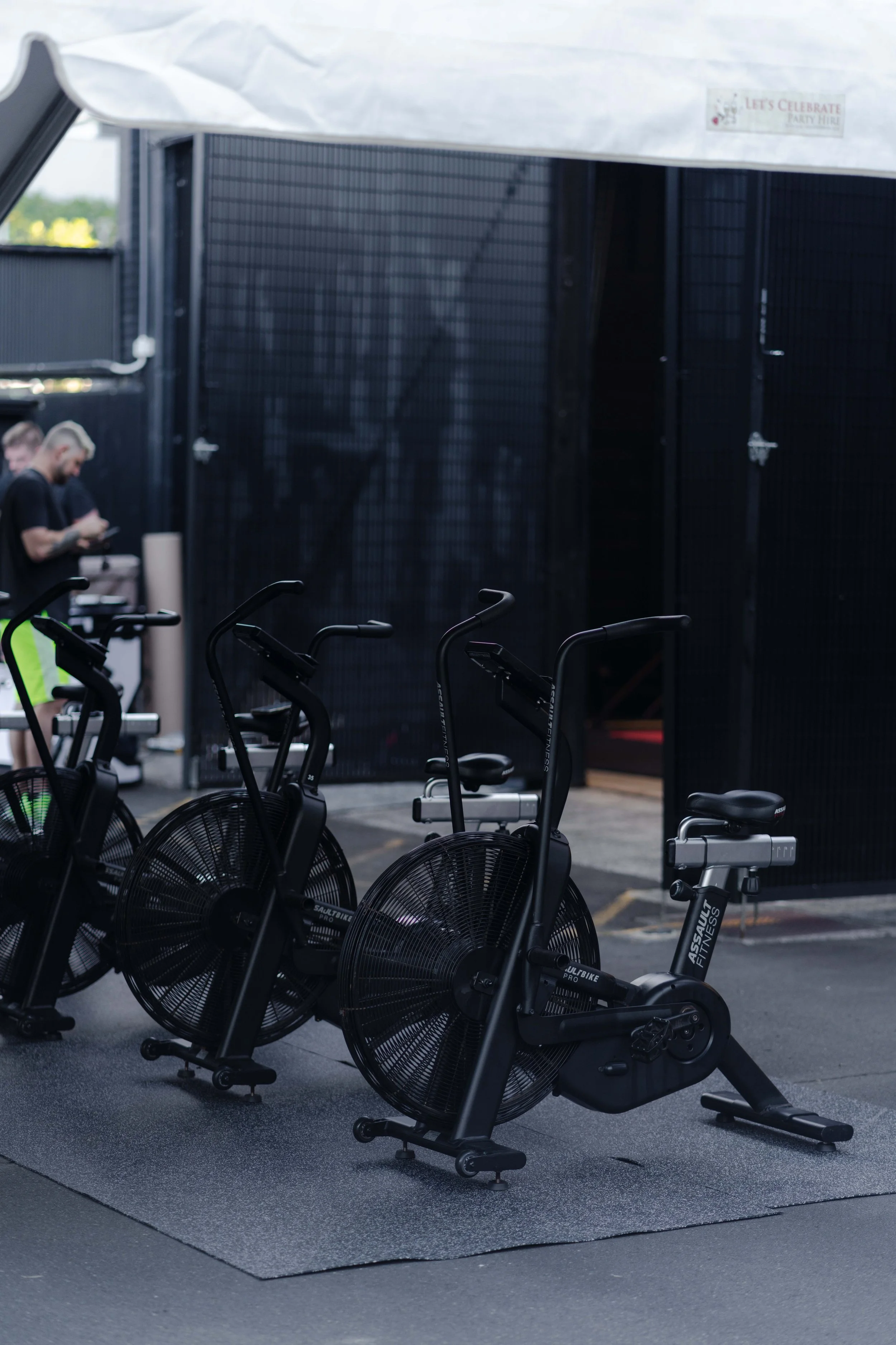 Three black stationary exercise bikes outdoors on a rubber mat, with black fencing and a small group of people in the background.