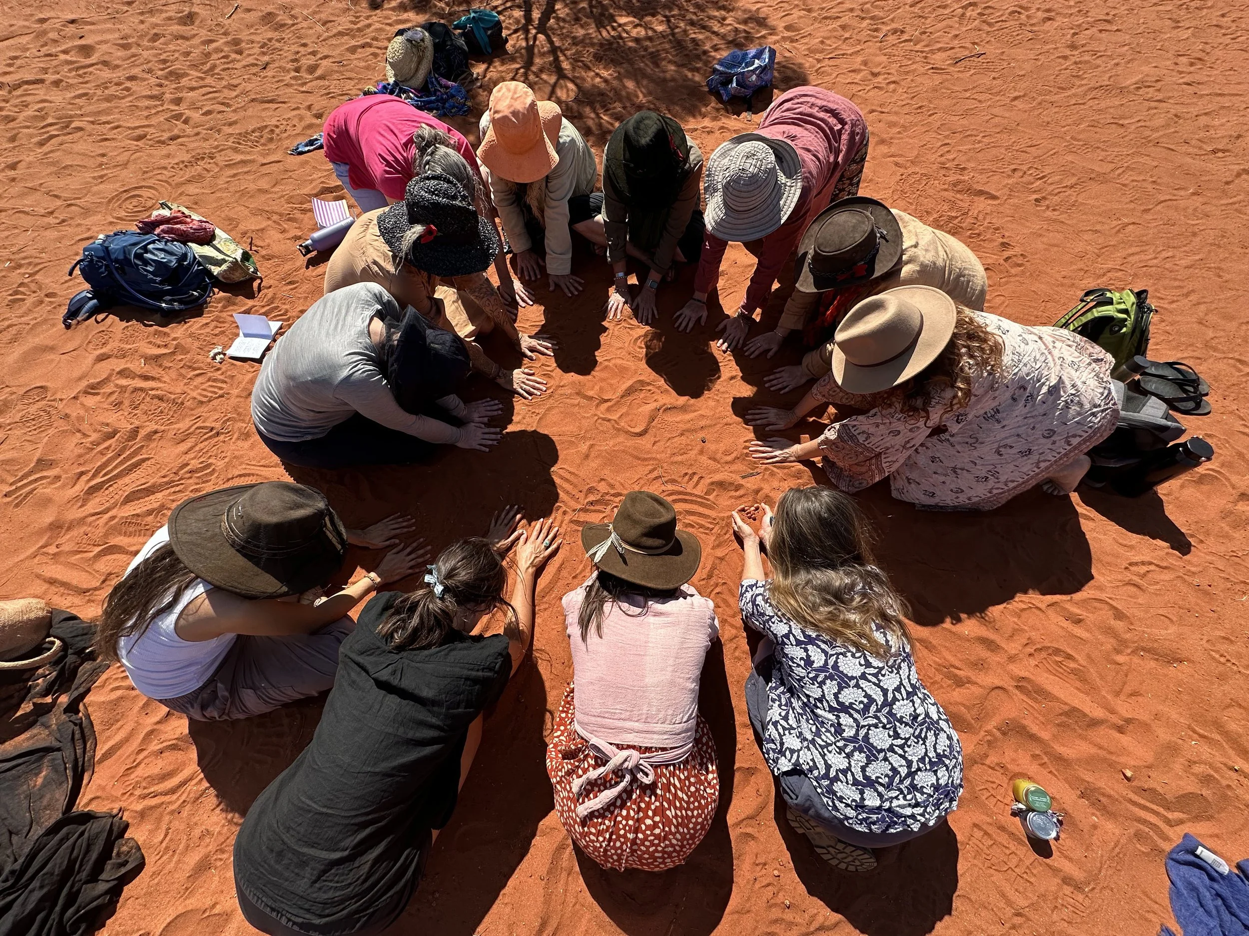 A group of people kneeling on the red desert sand, form a circular shape with their hands stretched forward, engaging in a group prayer or meditation.