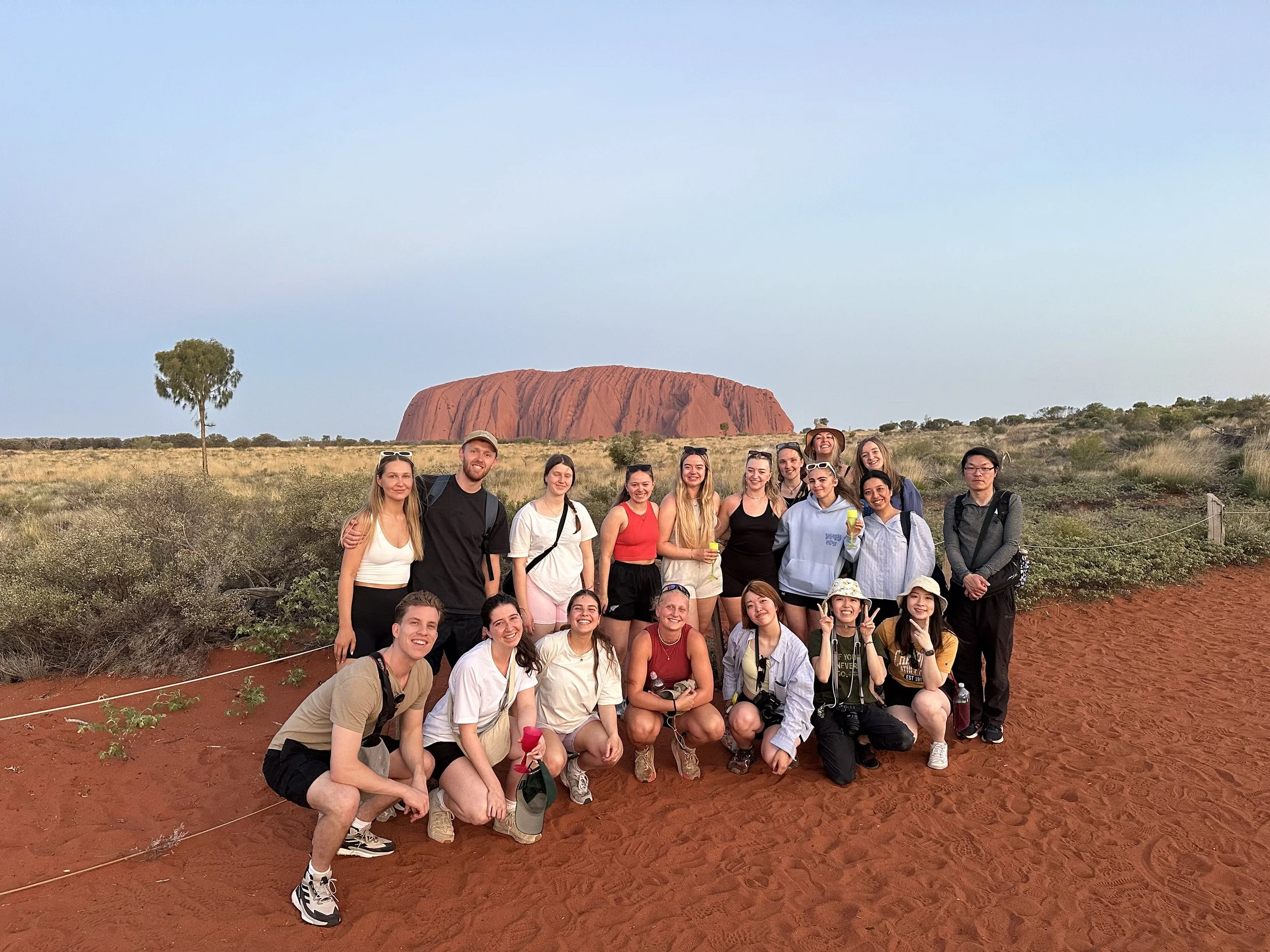 A group of twenty people posing for a photo in front of Uluru, a large red rock formation, in Australia during sunset.