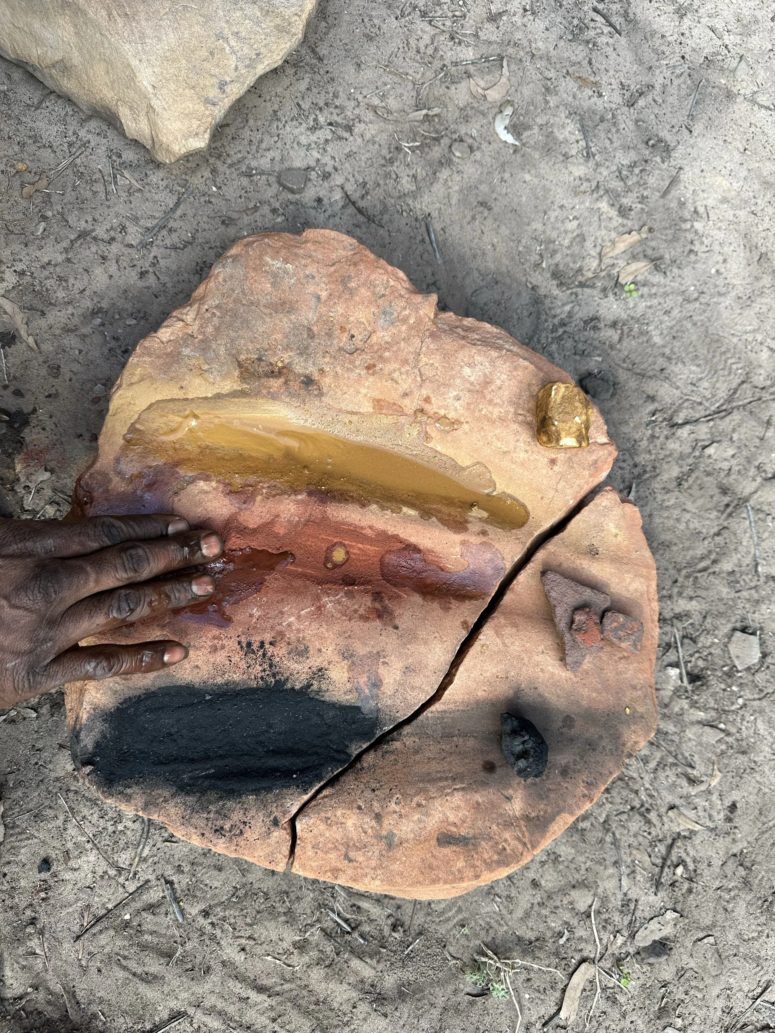 A large flat rock with various gold and black metallic objects embedded on its surface, placed on dirt with a person's hand resting on it.