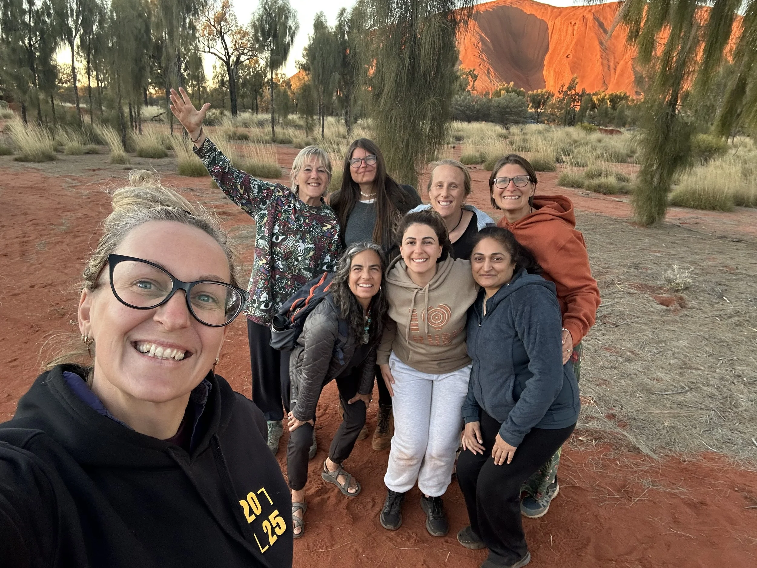 Group of nine women taking a selfie in a desert landscape at sunset, with red rocks and sparse vegetation in the background.