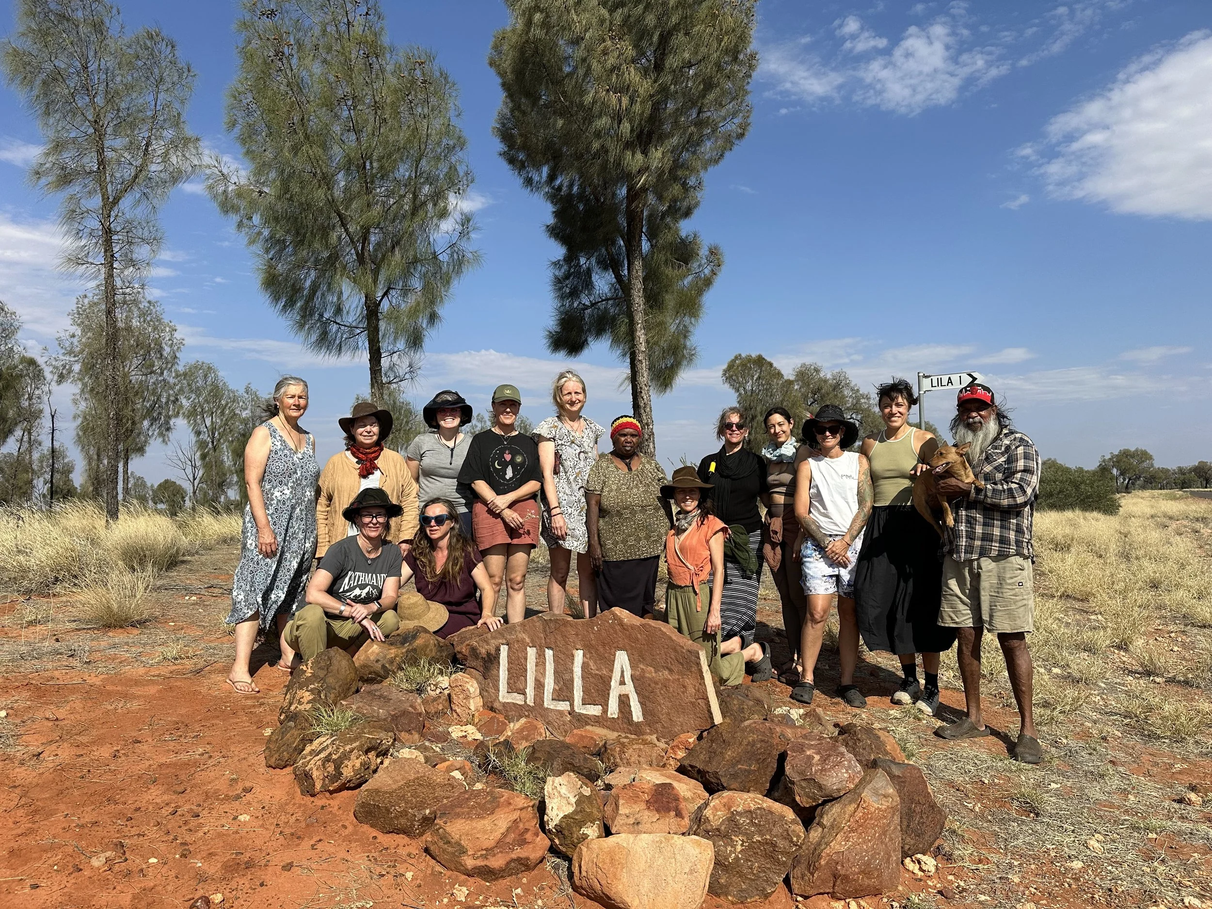 A group of approximately fifteen people standing outdoors in a natural setting with grass and trees. They are gathered around a large rock with the word 'LILLA' painted on it in white. The group is smiling, and some are wearing hats. The sky is blue 