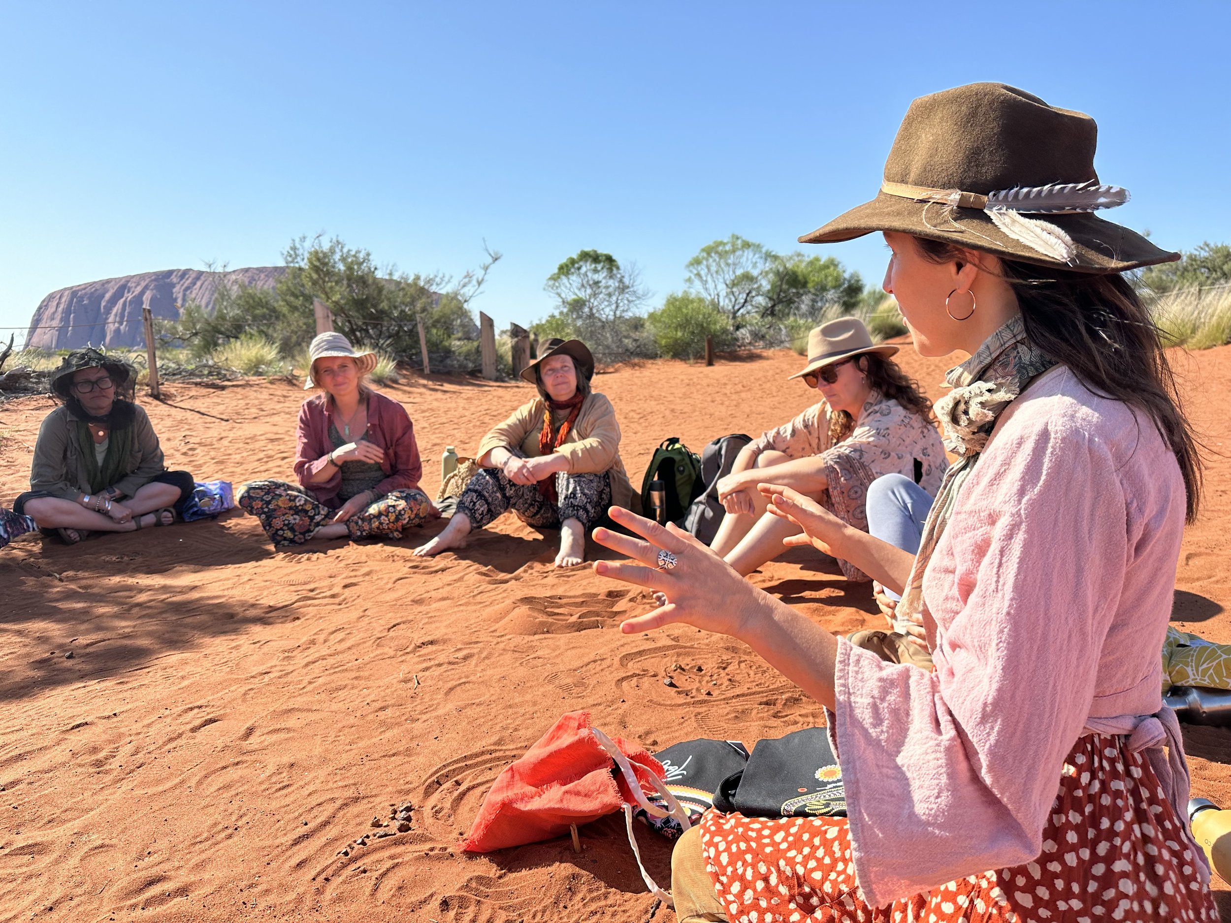 A group of women sitting on orange desert sand, wearing hats and casual clothing, participating in an outdoor discussion or gathering under a clear blue sky, with a large rock formation and sparse desert vegetation in the background.