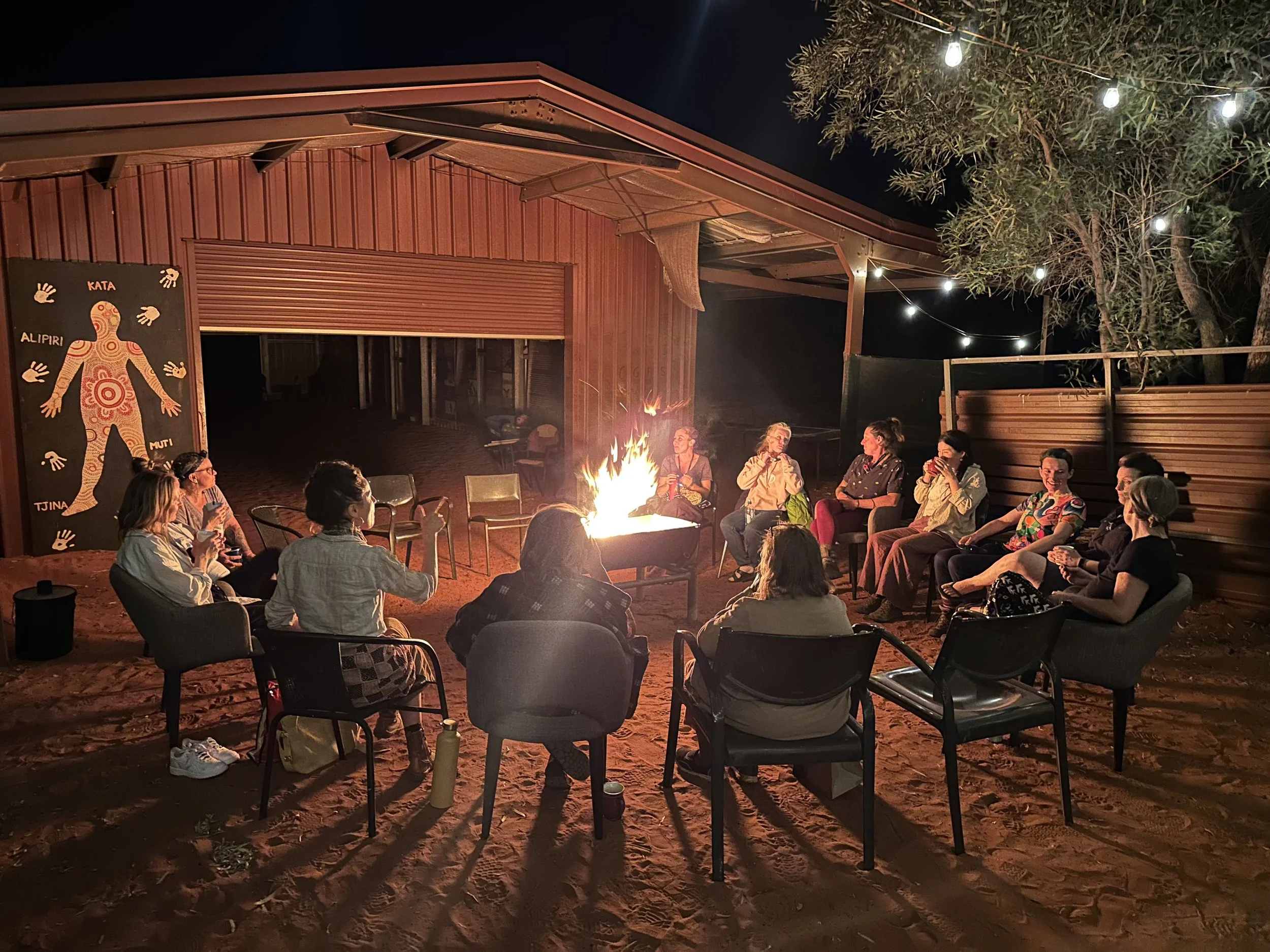 A group of people sitting around a campfire outdoors at night, with string lights overhead and a wooden building in the background.
