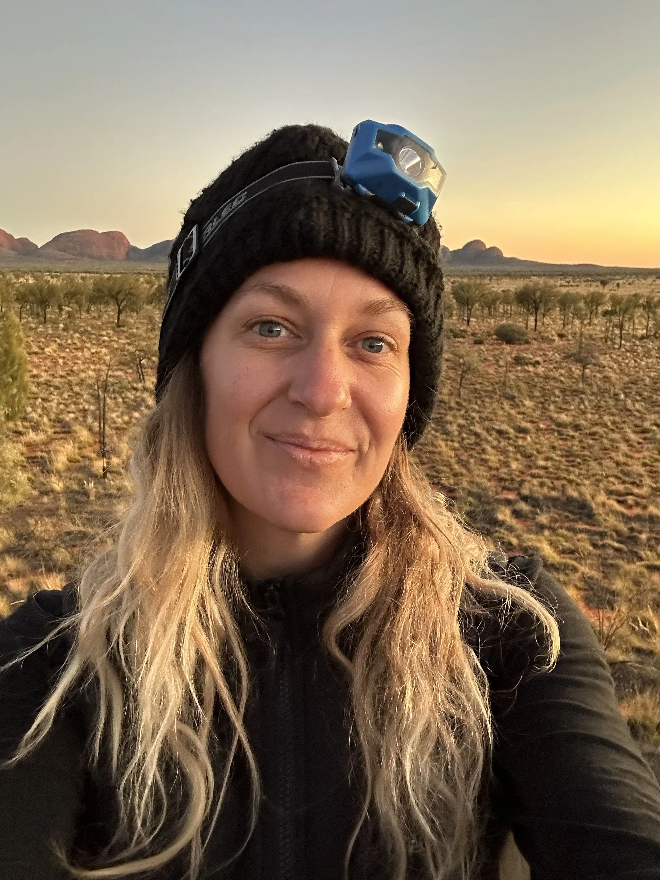 A woman taking a selfie outdoors in a desert landscape at sunset, wearing a black beanie with a headlamp.