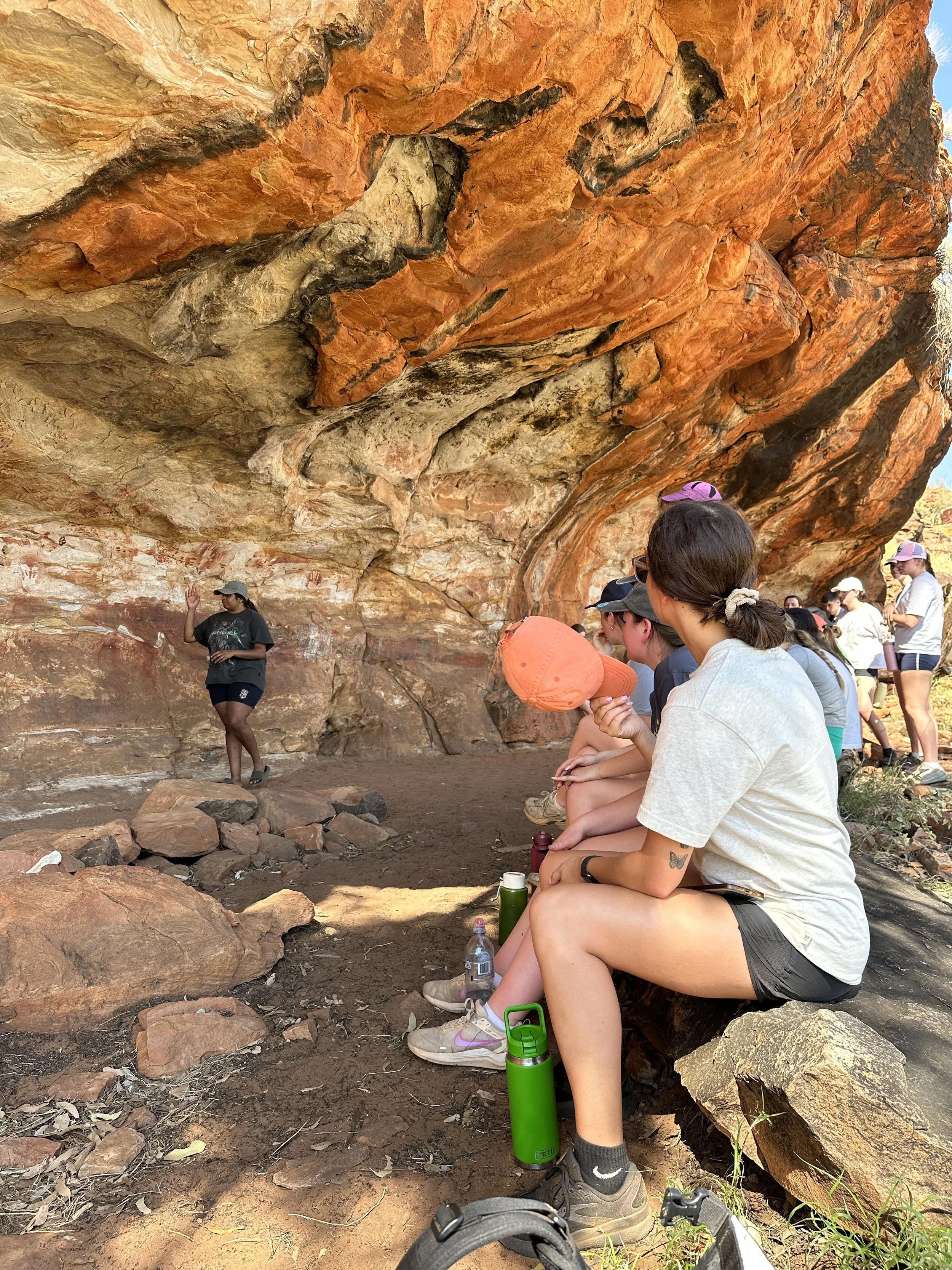 A group of people sitting on a bench under a large rocky overhang, watching a woman who appears to be giving a talk or demonstration outdoors in a rocky area.