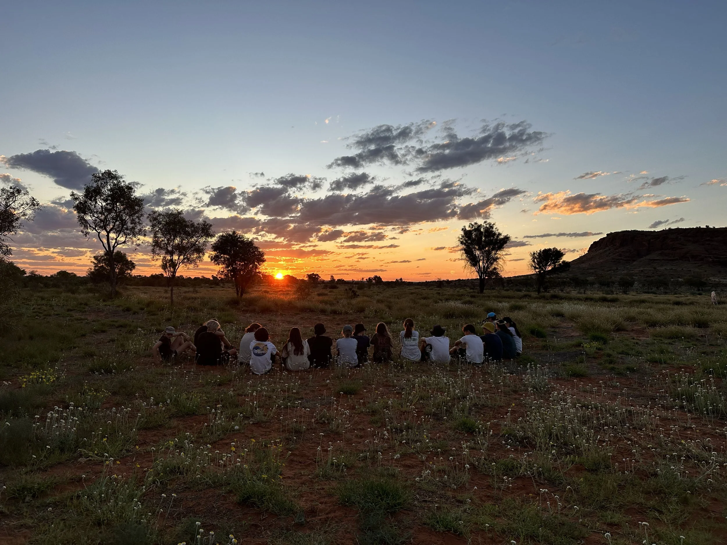 Group of people seated on the ground watching sunset in a grassy, semi-arid landscape with scattered trees and a distant hill.