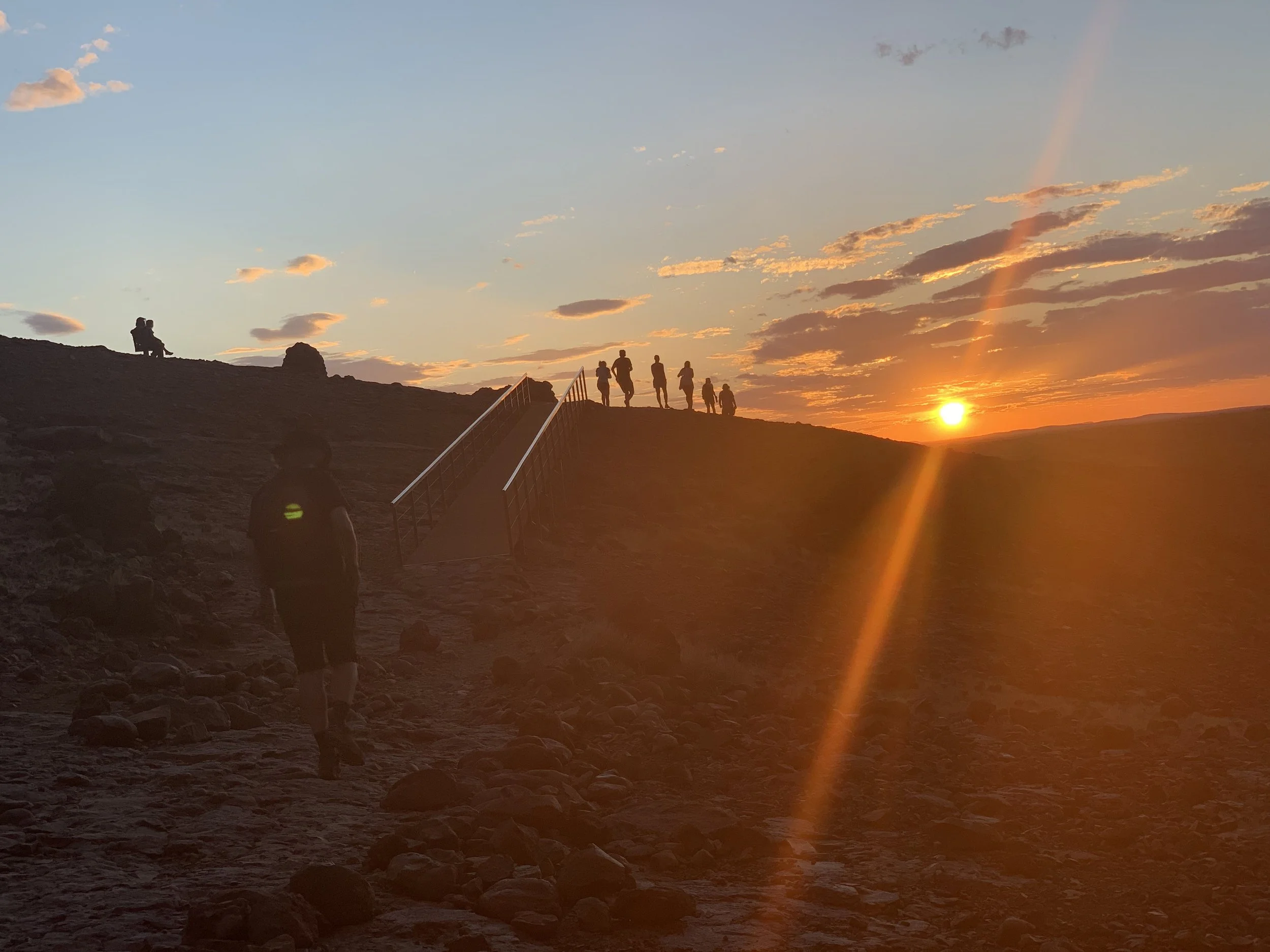 People walking along a trail during sunset with a hill on the right and a person sitting on a bench on the left.