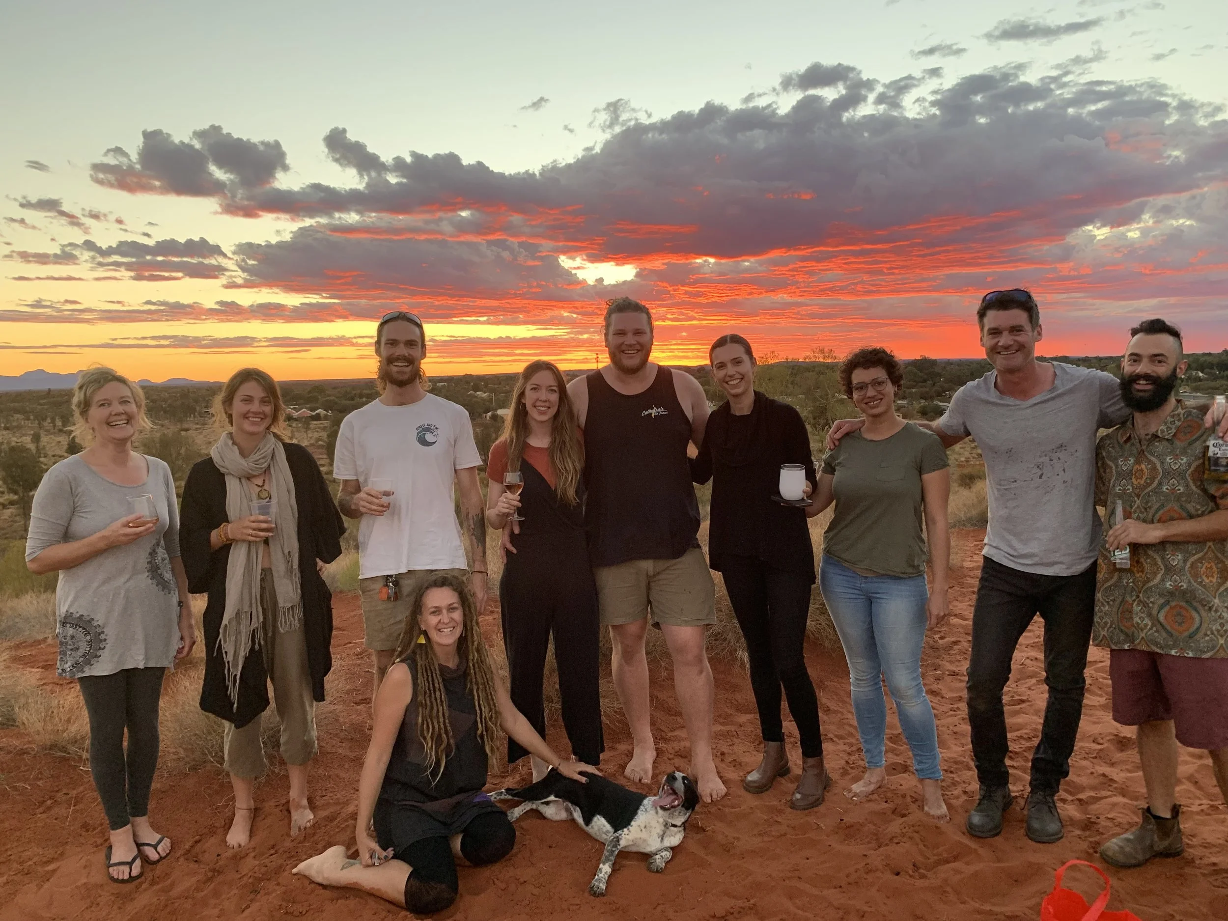 A group of ten people smiling and holding drinks at sunset in a desert landscape, with a dog sitting on the ground in front of them.