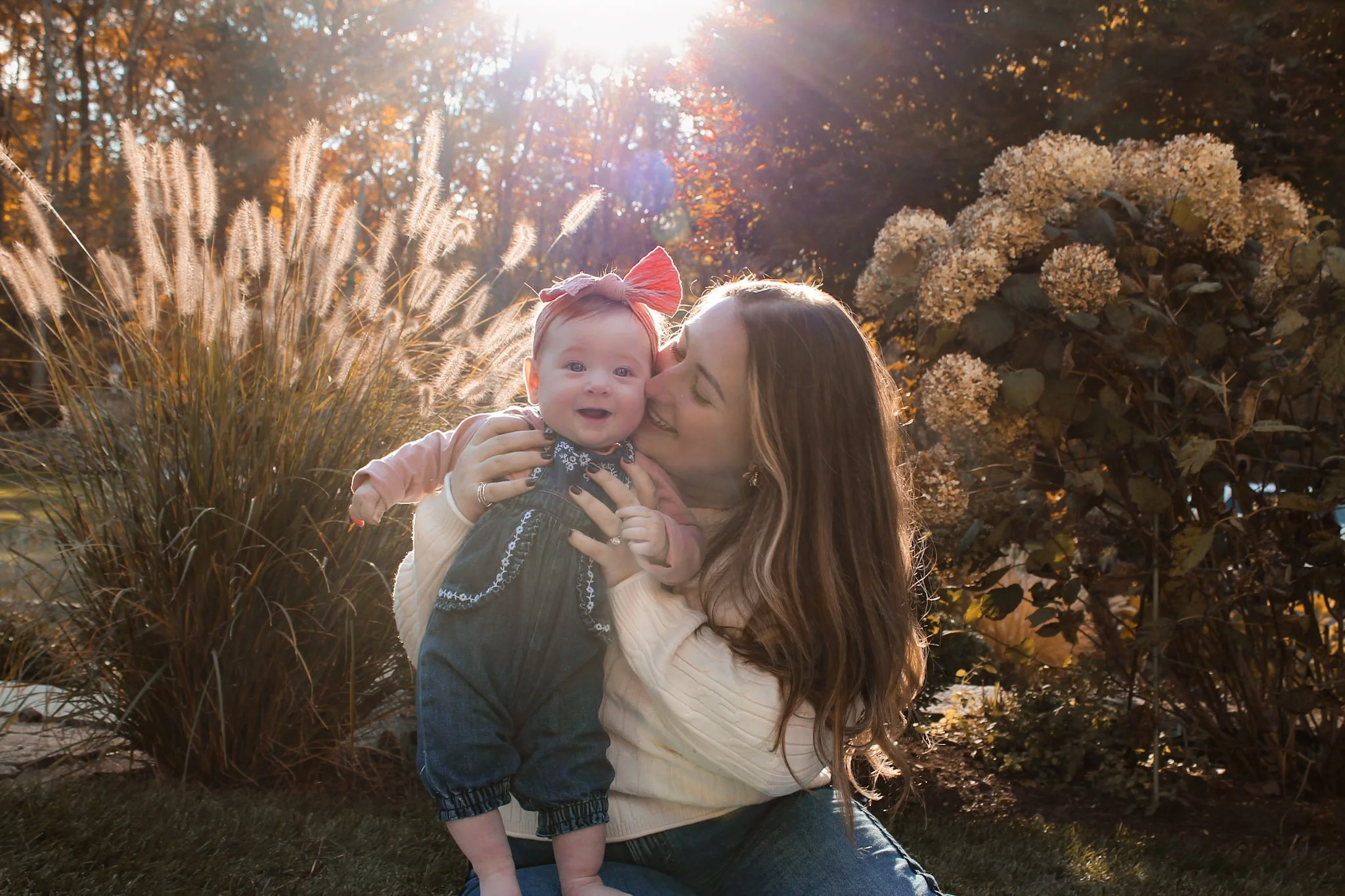 A woman holding a smiling baby girl outdoors during fall, with sunlight shining through trees and autumn foliage in the background.