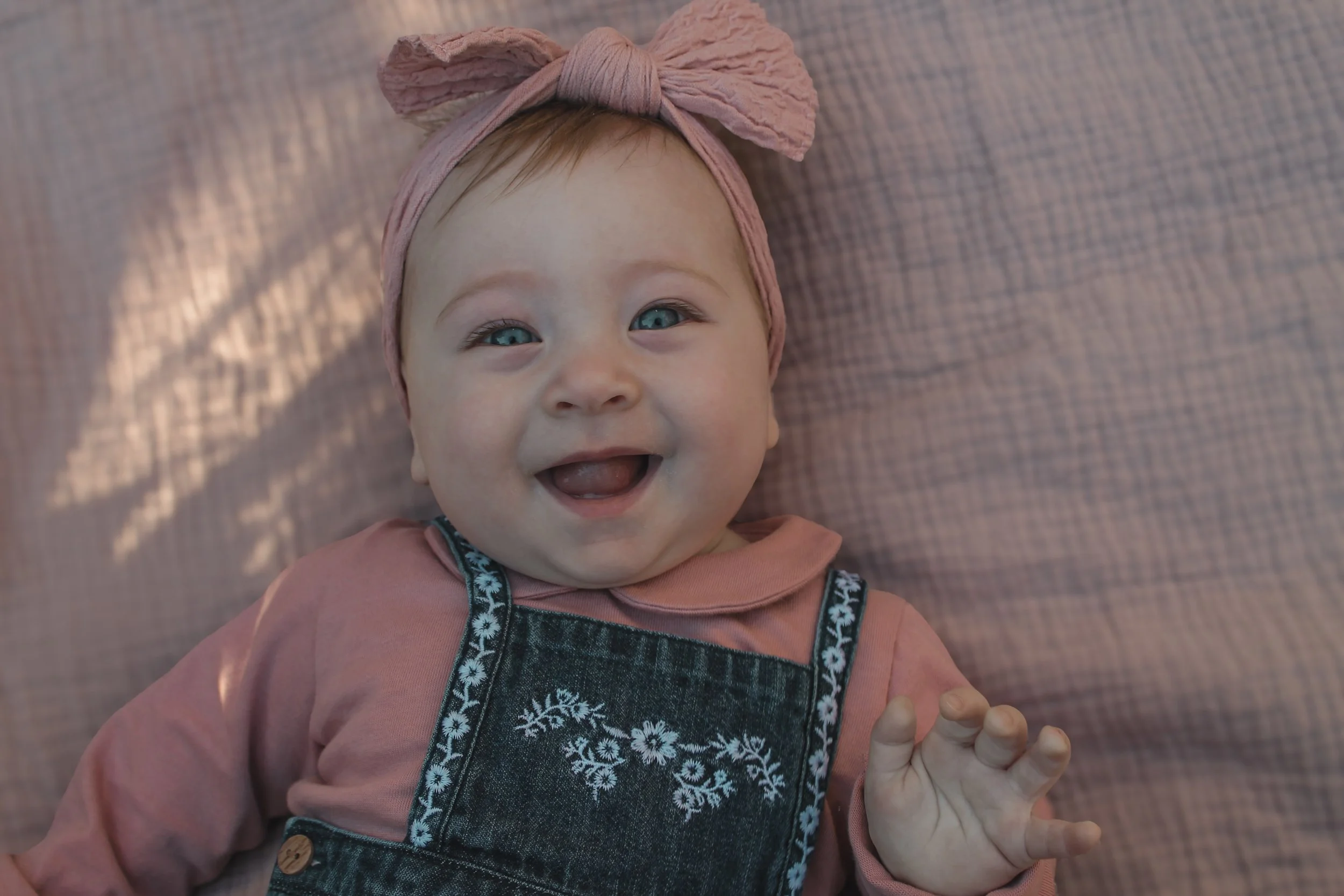 A smiling baby girl lying on a pink textured blanket, wearing a pink headband with a bow, a pink long-sleeve shirt, and denim overalls with embroidered floral patterns.