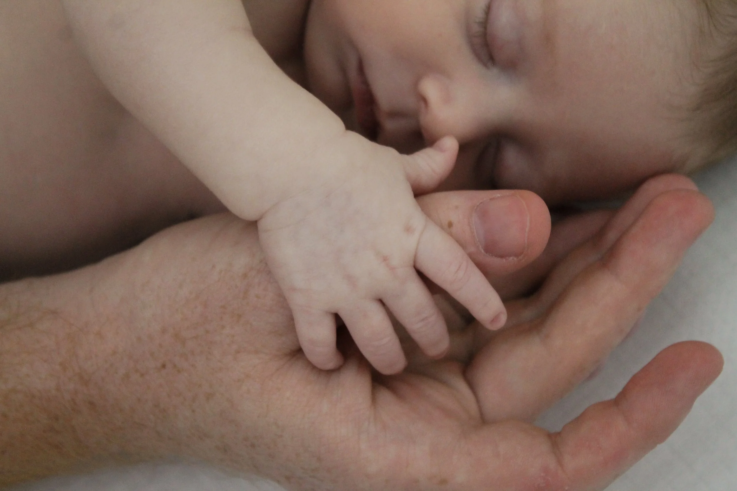 Close-up of a sleeping baby's face and hand gently holding an adult's finger.