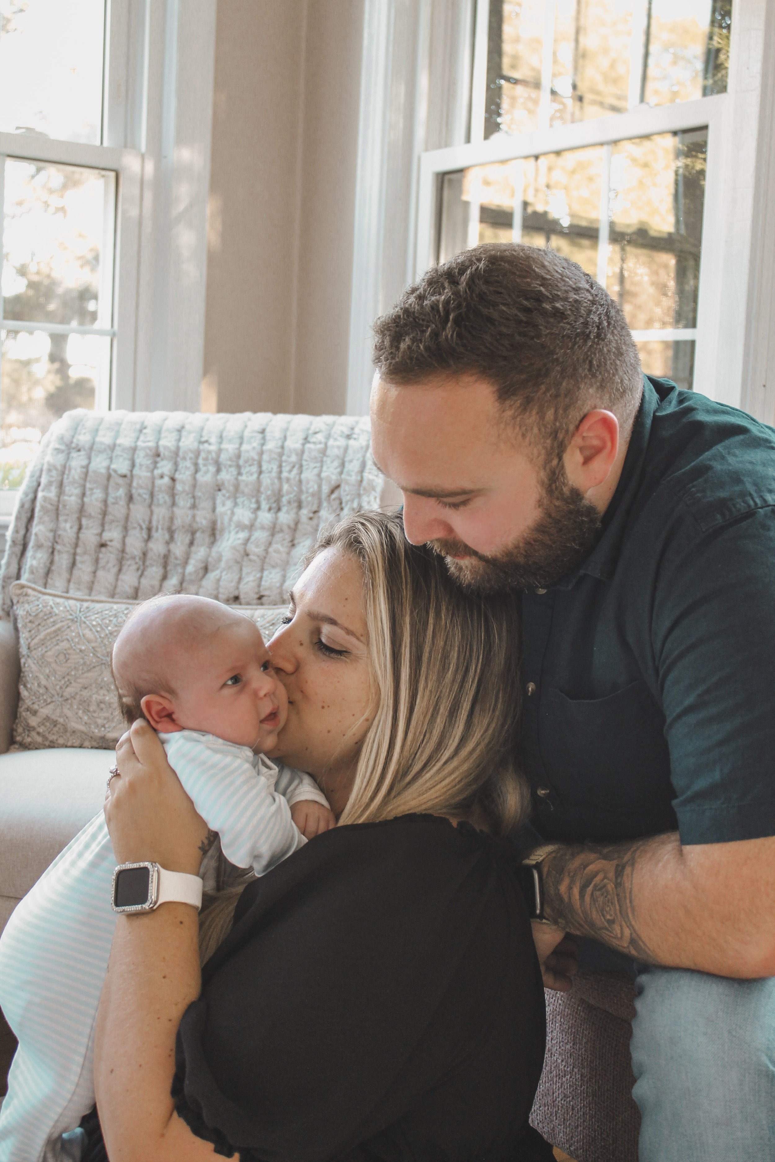 A woman holding a newborn baby while a man kisses her on the forehead inside a room with large windows and beige walls.