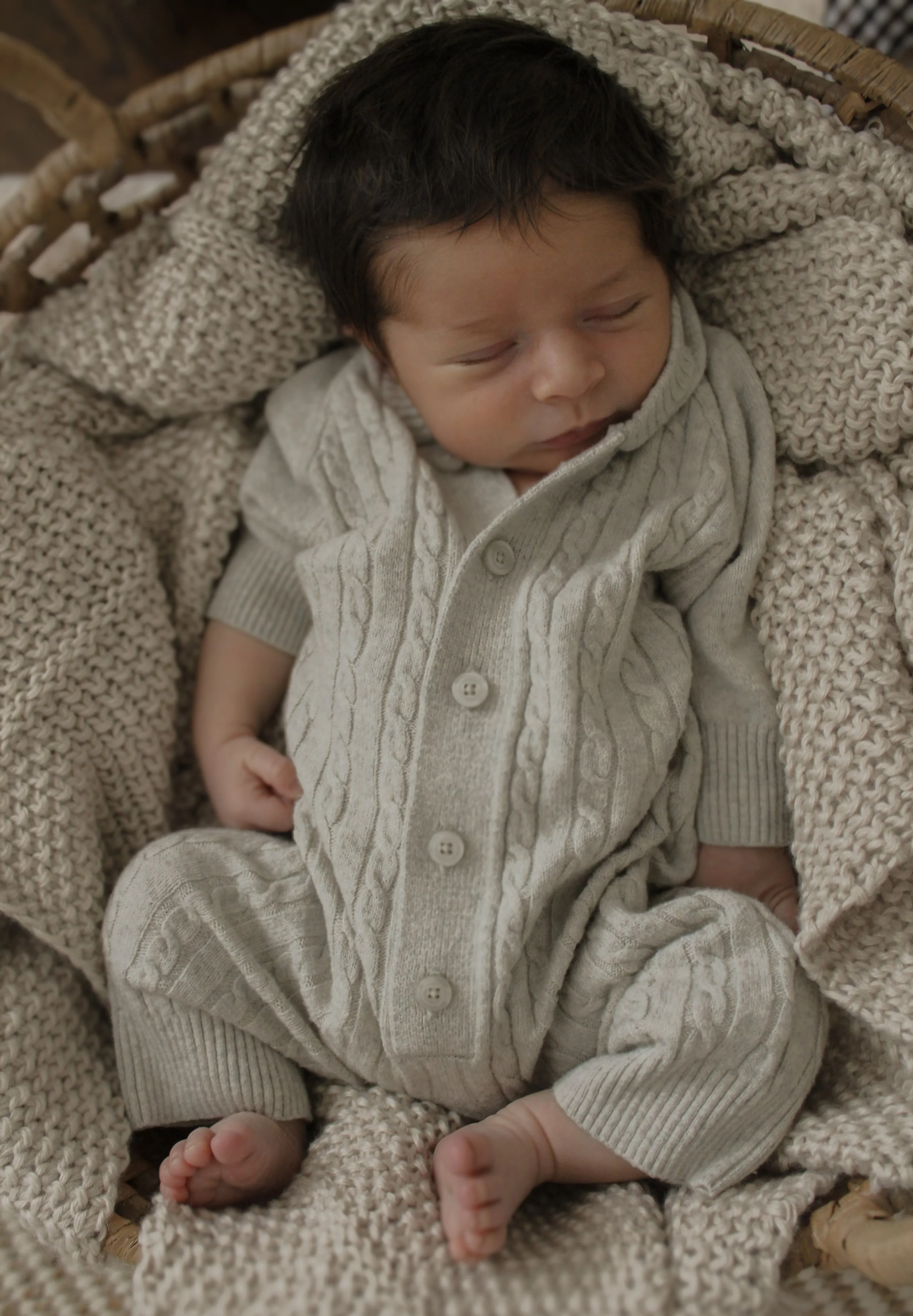 A sleeping baby with dark hair, dressed in a light gray, cable-knit patterned onesie, laid in a cozy wicker basket surrounded by a chunky knit blanket and a knitted blanket.