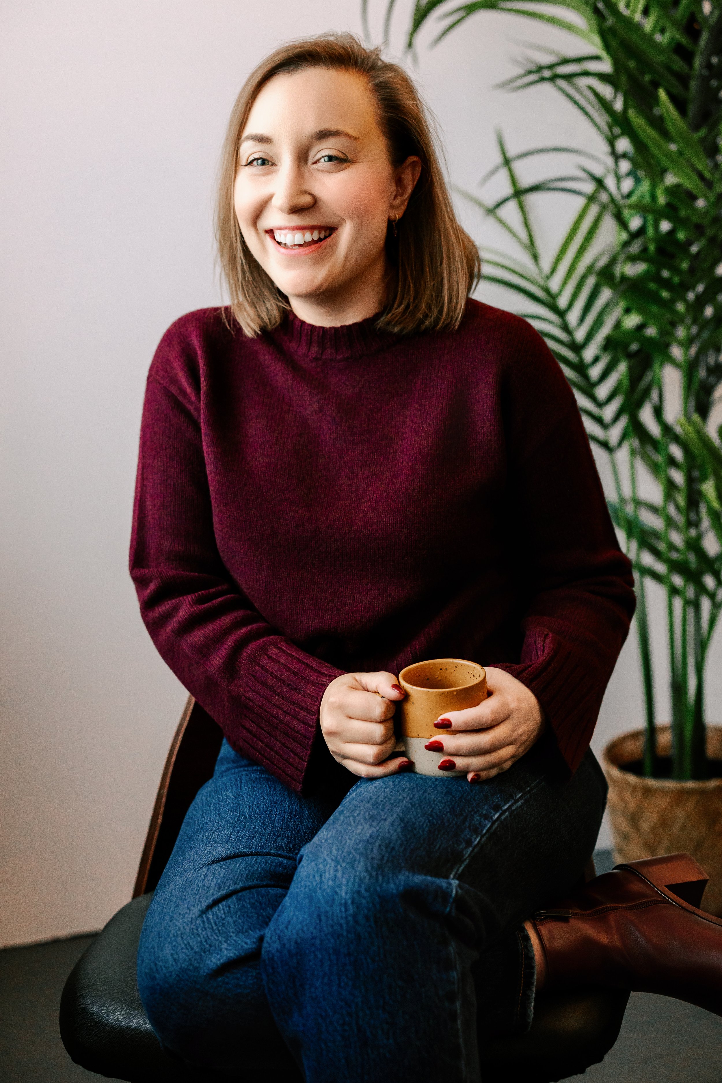 A woman with short brown hair and light skin, smiling and sitting on a black chair, holding a beige ceramic mug with both hands. She is wearing a maroon sweater, blue jeans, and brown boots, with a large green potted plant in the background.