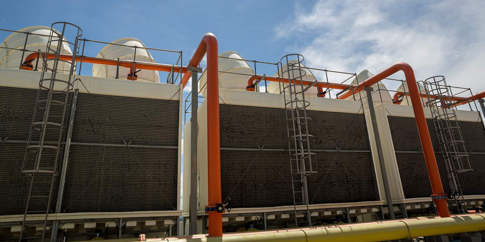 Industrial equipment with large black panels, orange pipes, ladders, and vents against a cloudy sky.