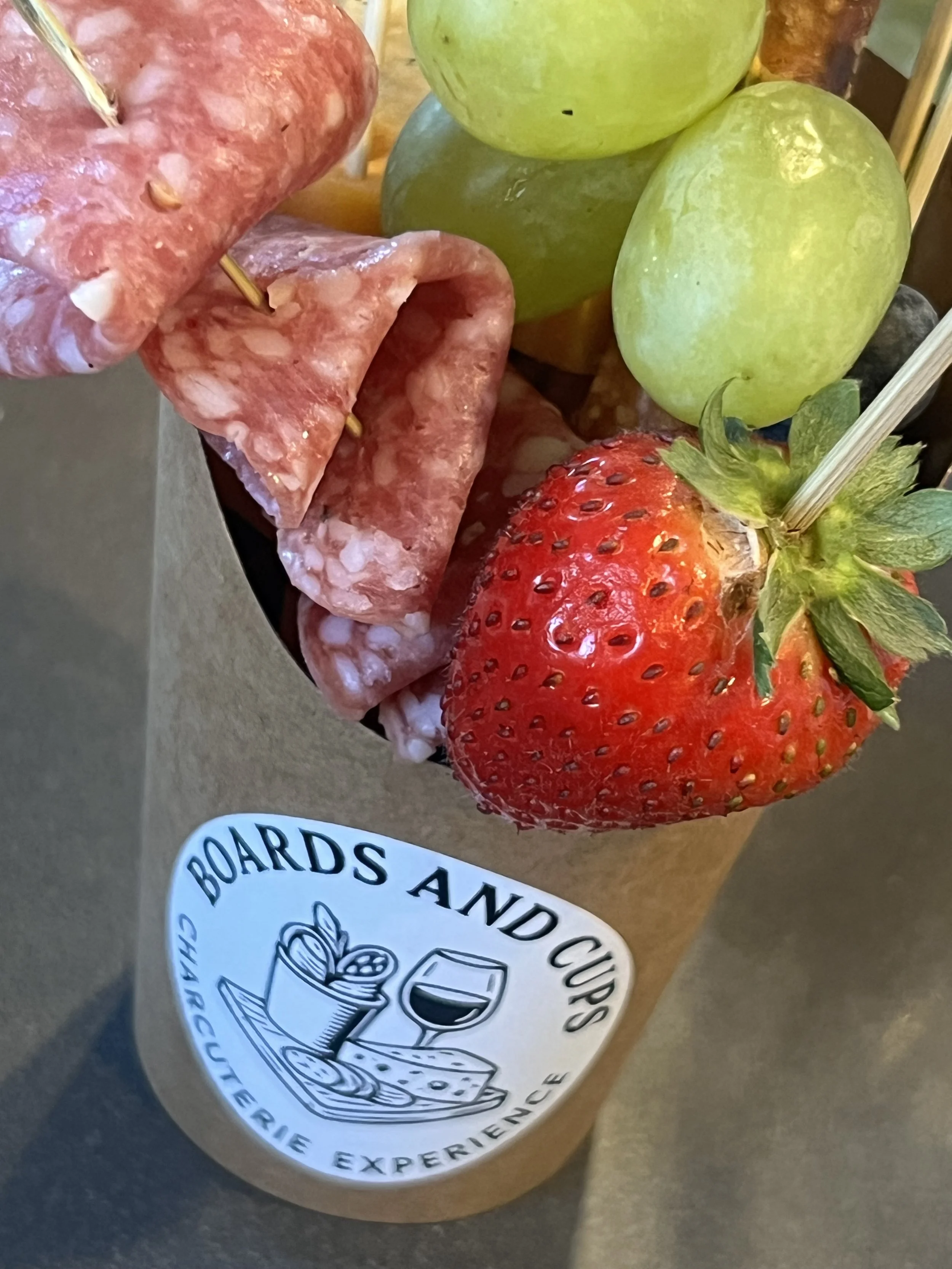 Fruit and charcuterie skewer with grapes, strawberry, and salami, in a cup labeled "Boards and Cups"